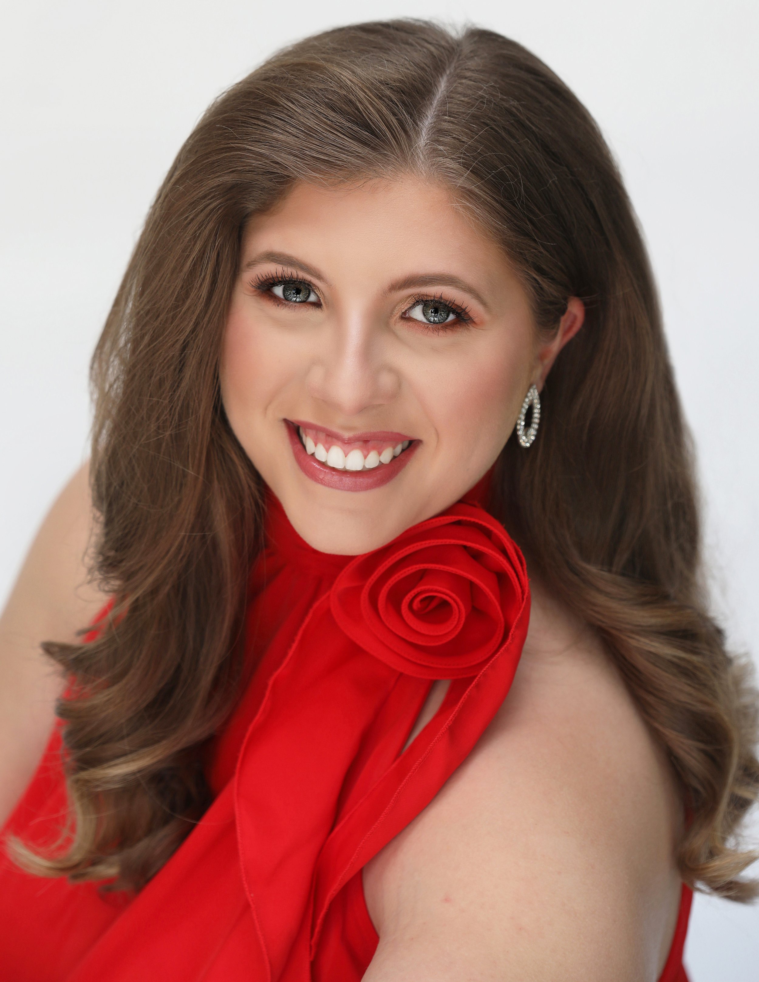 A woman with long, wavy brown hair and bright blue eyes, smiling widely in a red dress with a floral detail, against a white background.
