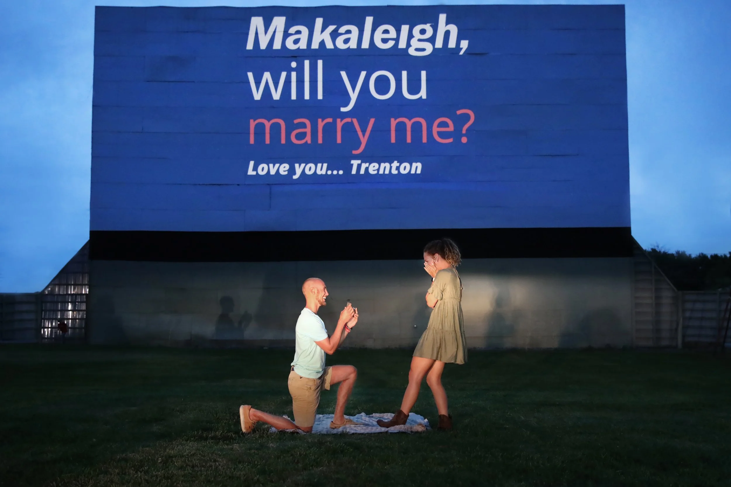 A man proposes marriage to a woman on a grassy outdoor area during dusk, with a large billboard in the background displaying a proposal message.
