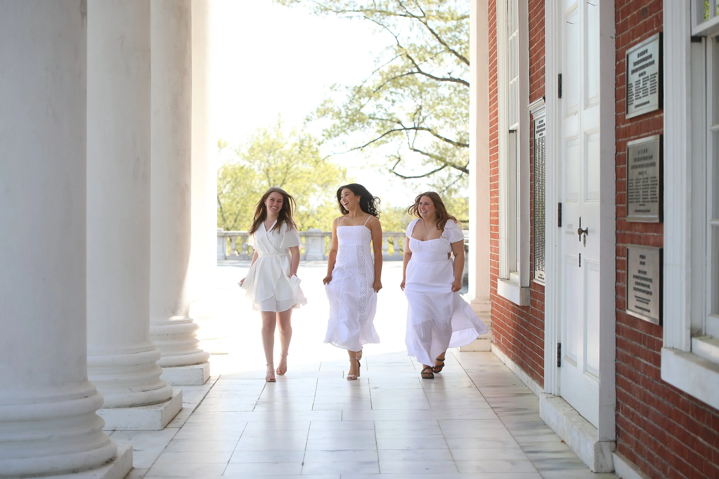 Three women walking and smiling in a sunlit outdoor corridor with white columns and brick walls, dressed in white summer dresses.