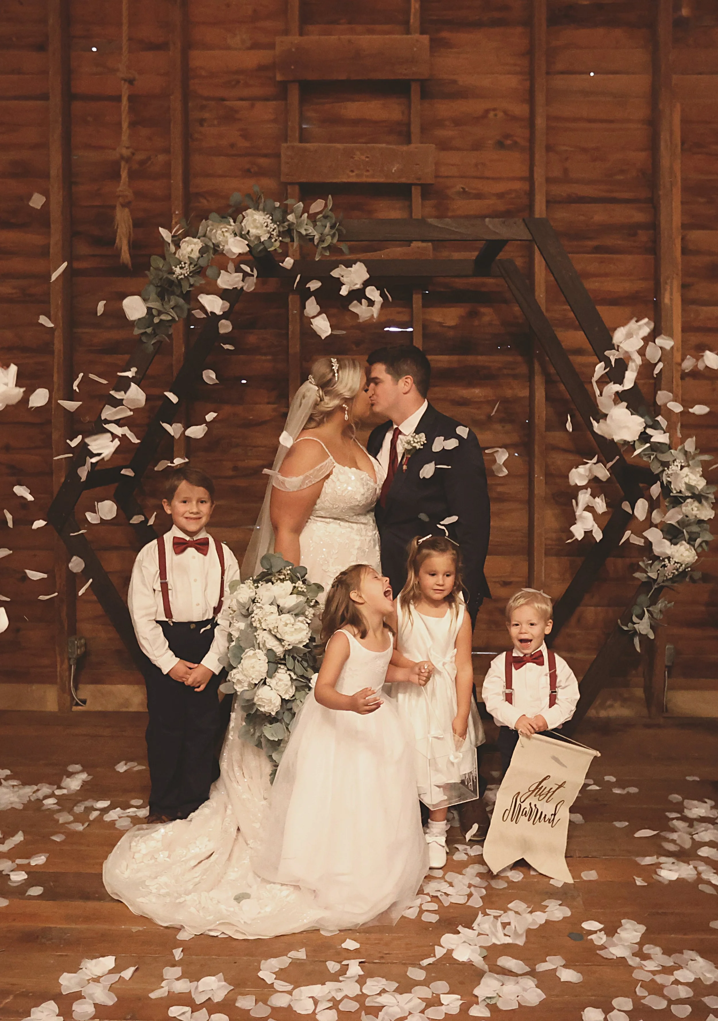 A bride and groom kissing at their wedding ceremony, surrounded by children and rose petals, in a rustic wooden setting.