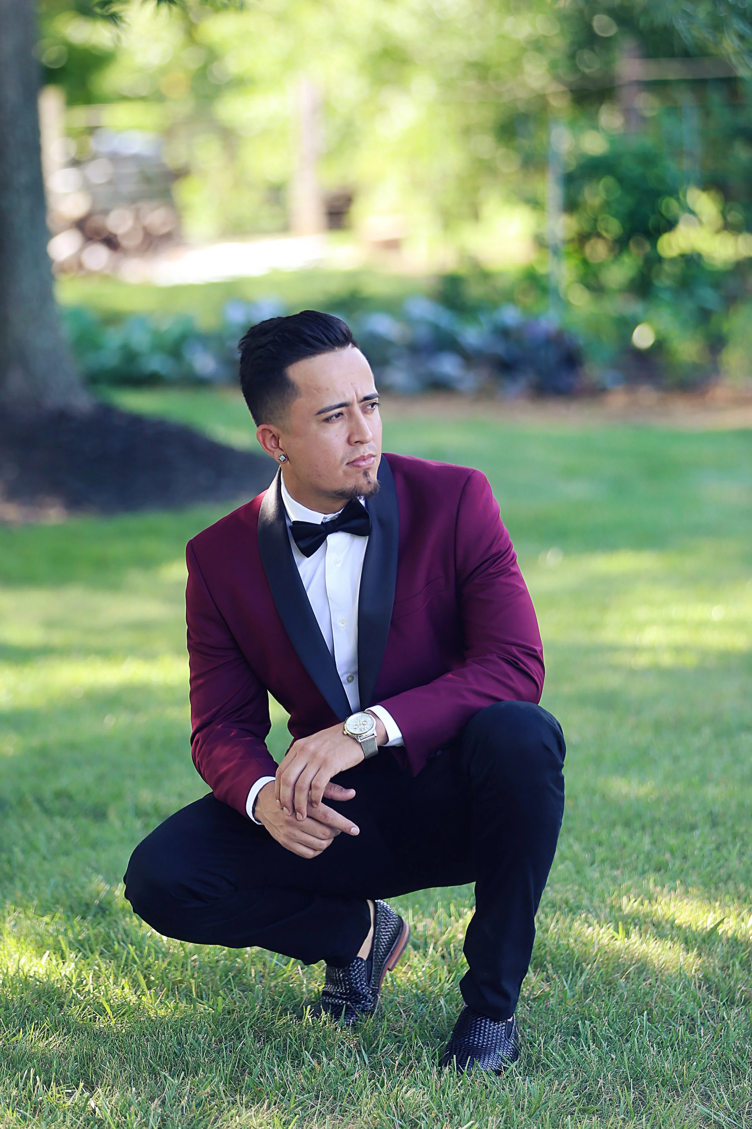A man in a tuxedo with a maroon jacket, black lapel, white shirt, and black bow tie, crouching on grass outdoors with trees and blurred background.