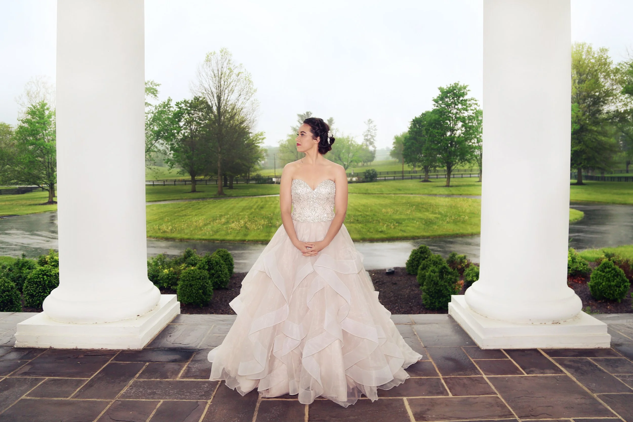 A woman in a strapless white wedding gown stands on a porch between two large white columns, with a lush green landscape and trees in the background.
