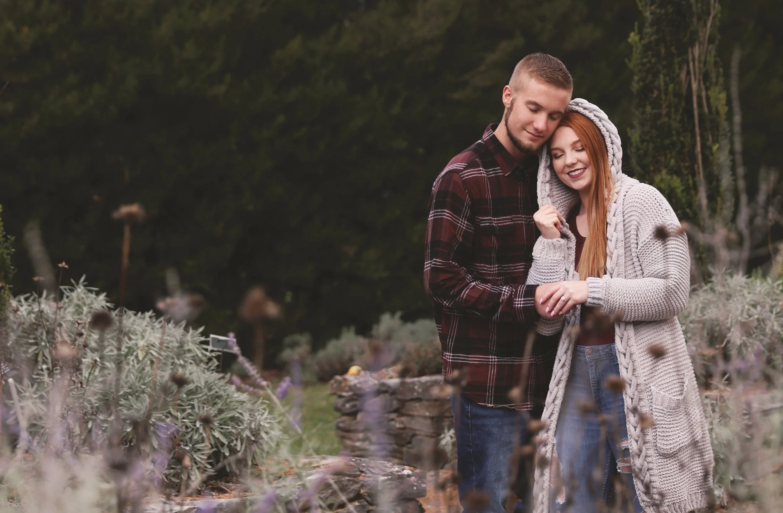 A young couple embracing outdoors on a fall day, surrounded by plants and a stone wall, with trees in the background.