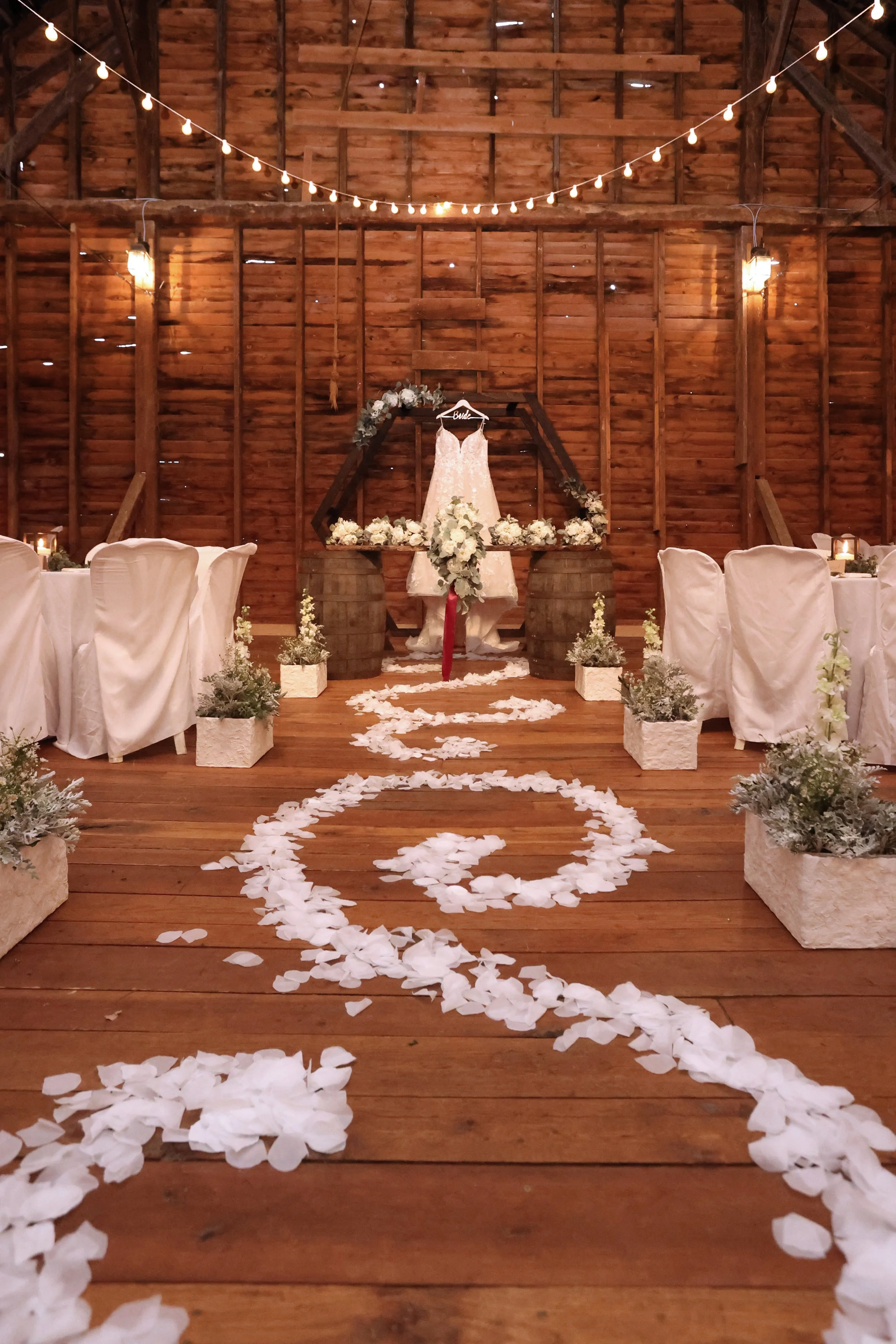 Wedding aisle decorated with white flower petals leading to an altar with a wedding dress hanging on a black wooden frame, surrounded by floral arrangements and rustic wooden barrels, inside a barn with string lights overhead.