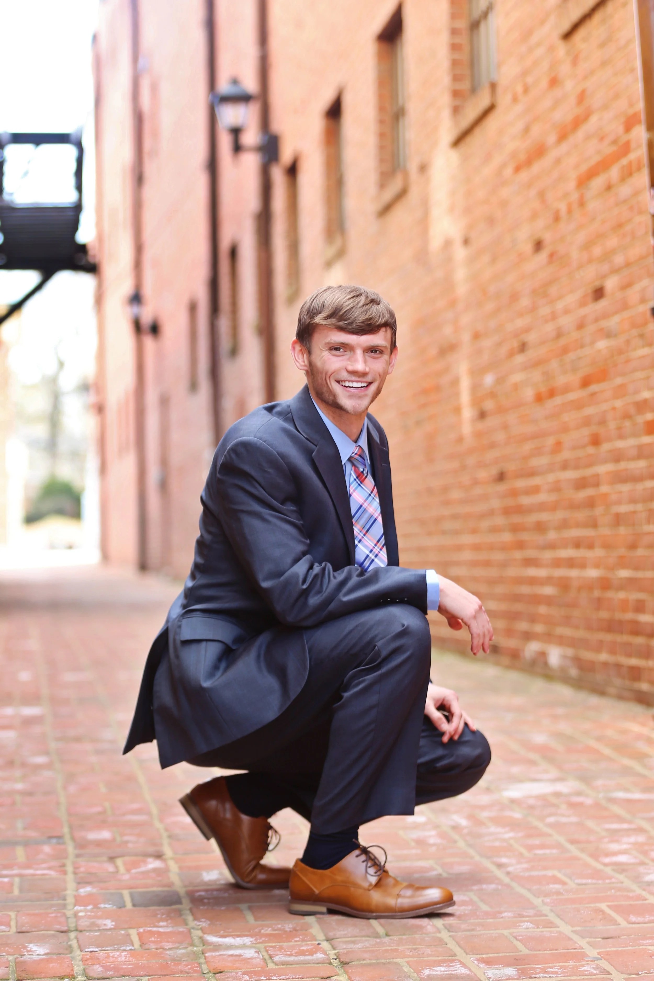A young man in a business suit crouching down, smiling, on a brick alleyway with brick buildings and outdoor lamps