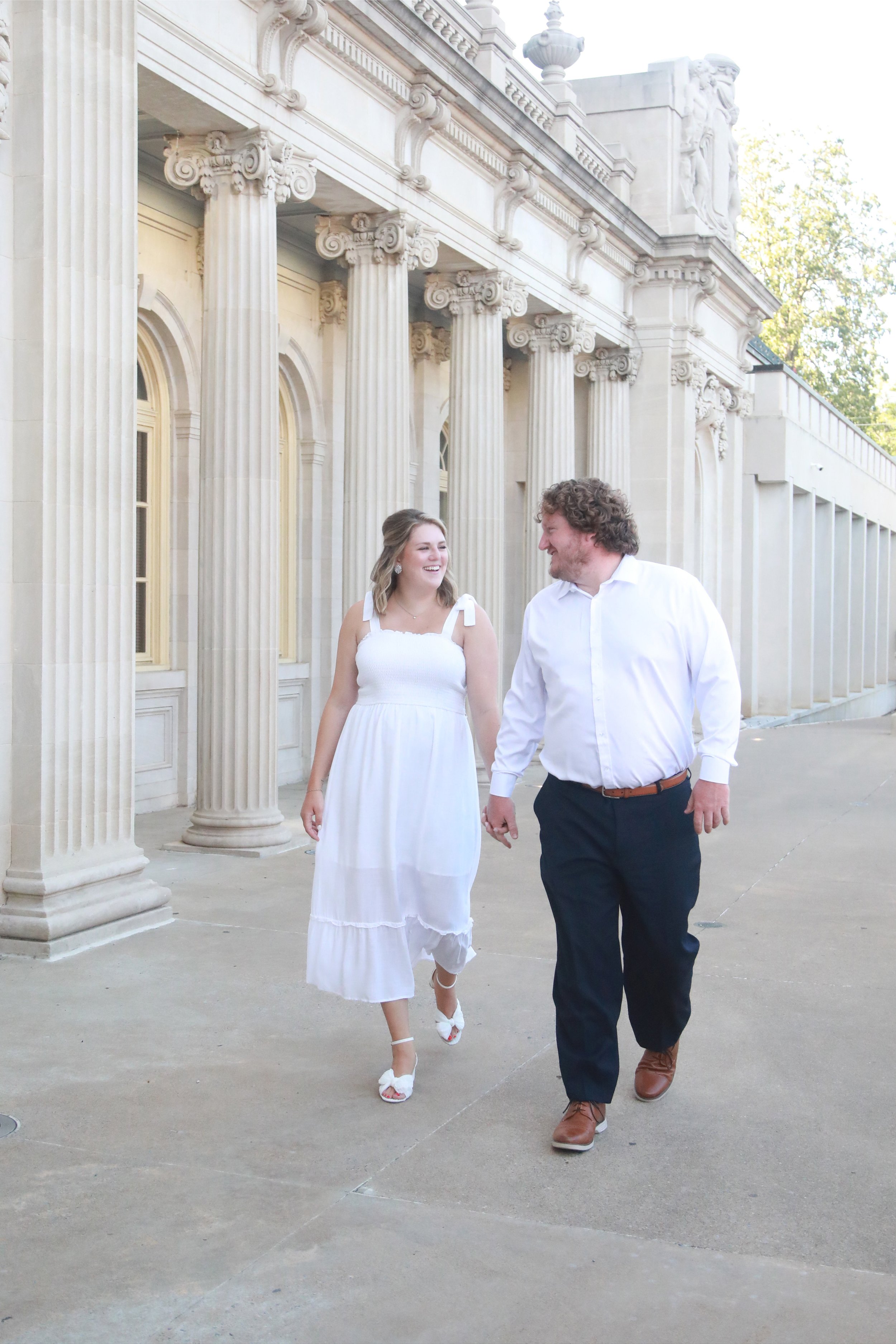 A woman and a man are walking hand in hand outside a building with tall, classical columns and ornate architectural details, smiling at each other.