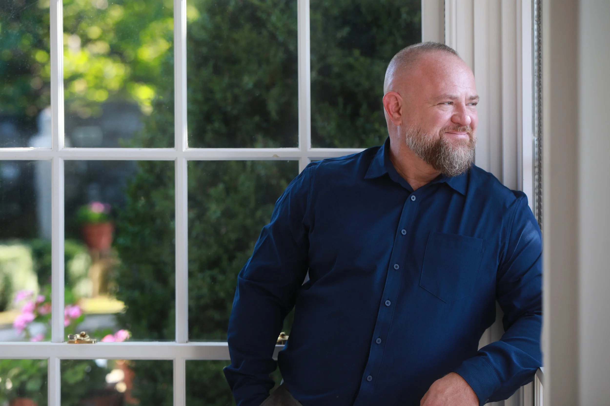 A man with a beard and short hair, wearing a navy blue button-up shirt, stands by a window looking outside with a slight smile.