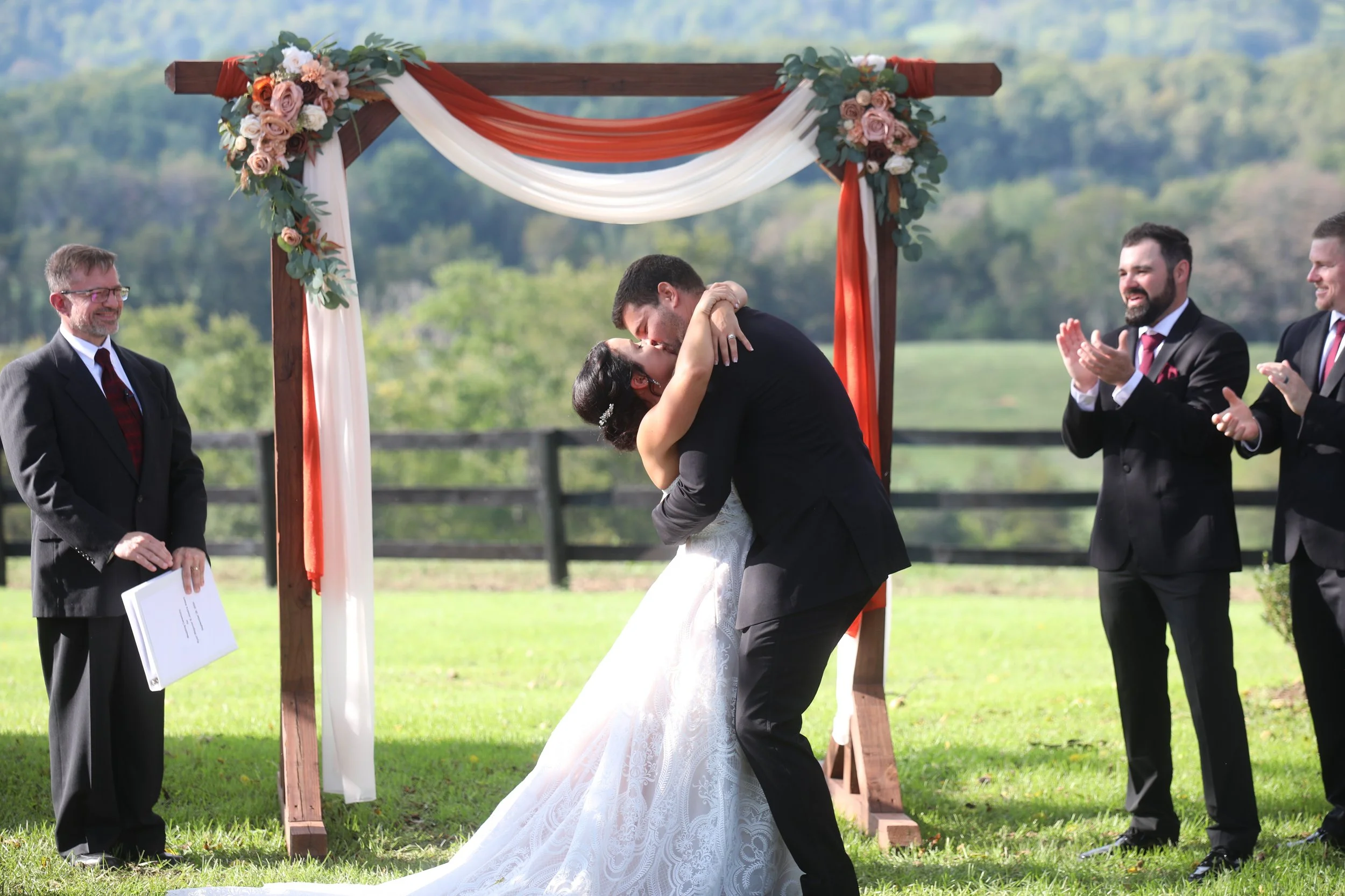 A bride and groom share a kiss during an outdoor wedding ceremony, under a decorated wooden arch, with two men in suits clapping and smiling around them.