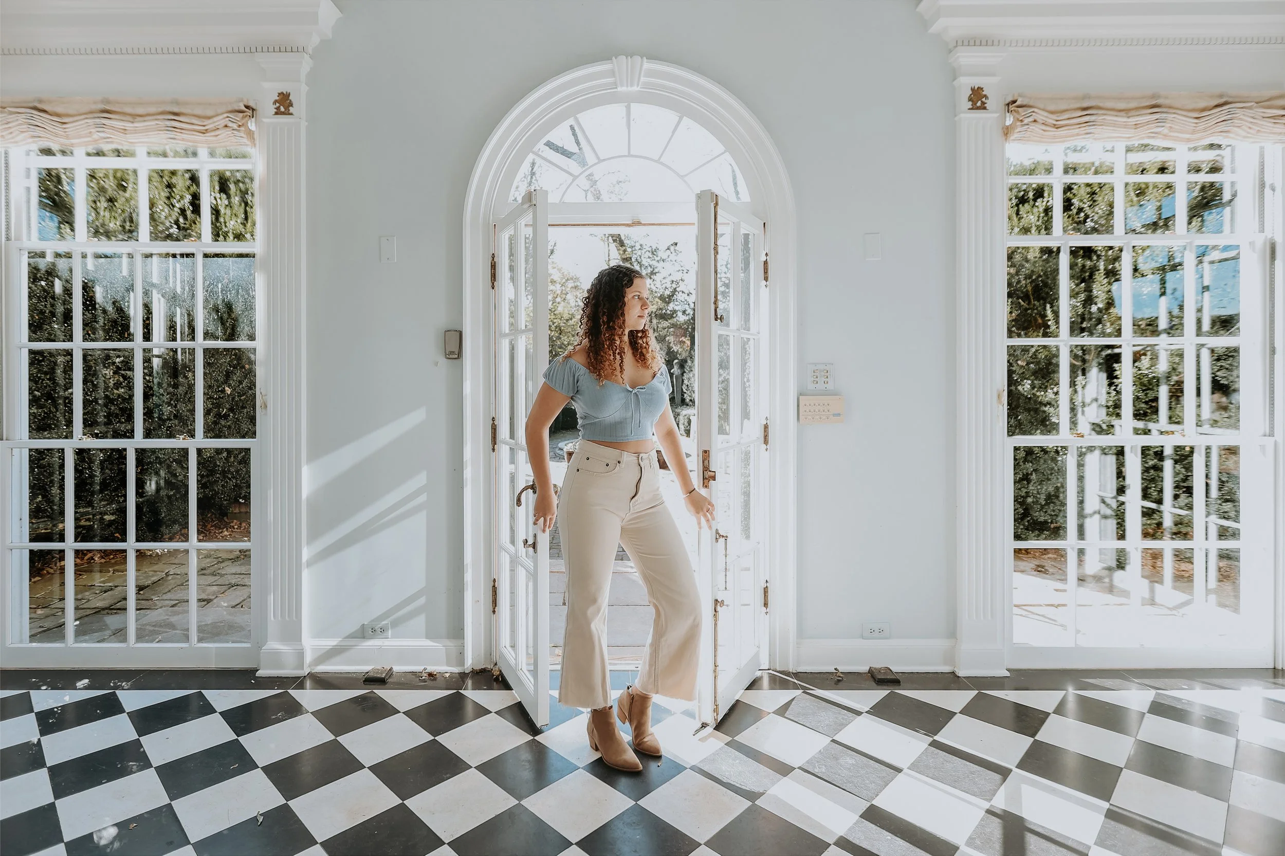 Young woman standing in front of open glass door in a bright, white-room with large windows and checkered floor.