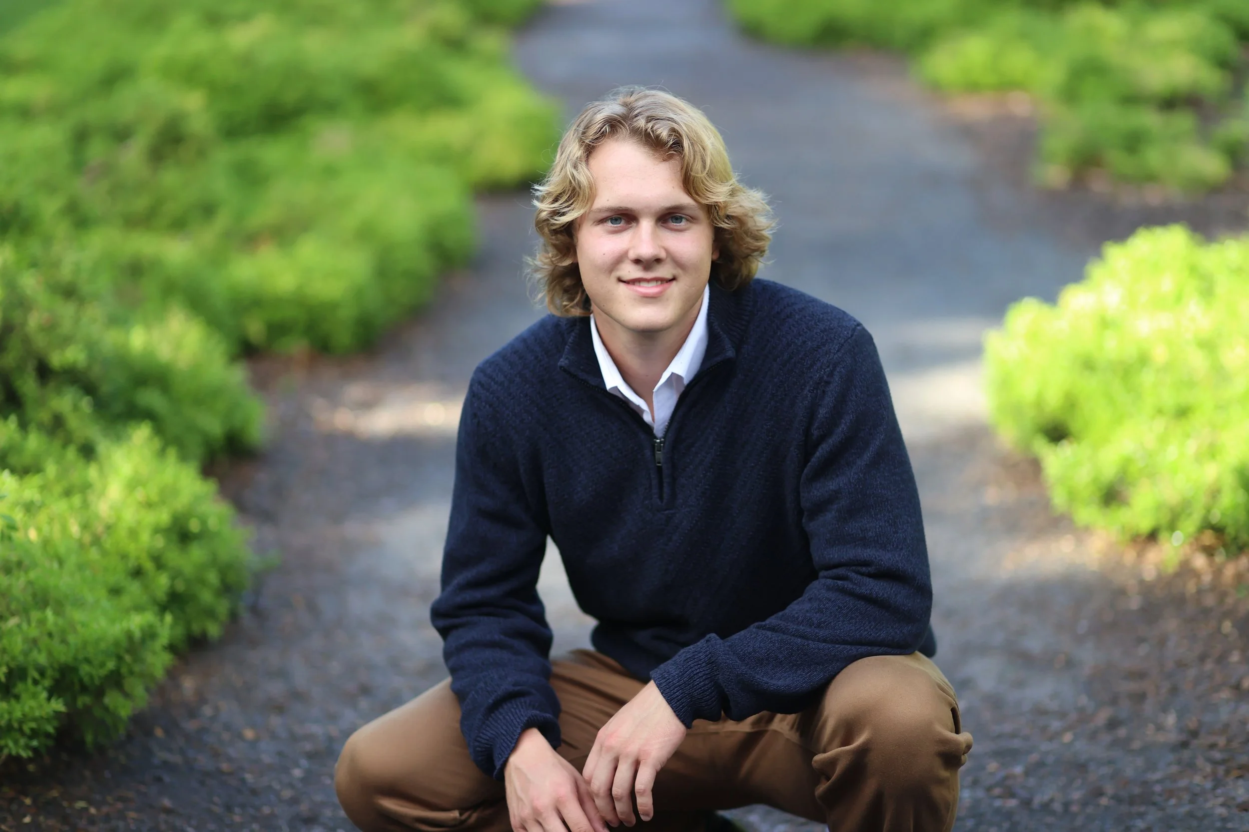 A young man with blonde, wavy hair, wearing a navy sweater and khaki pants, squatting on a gravel path surrounded by greenery.