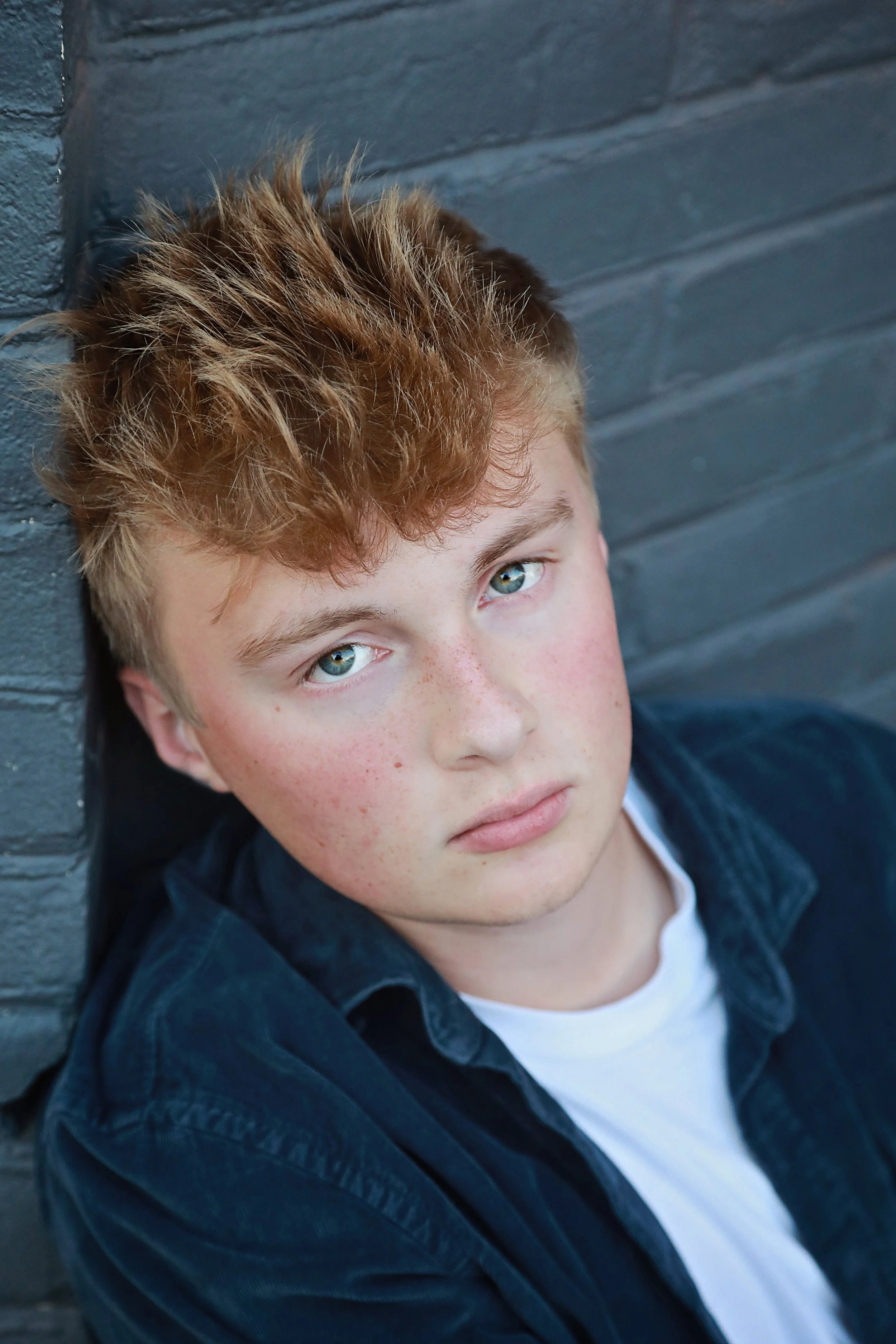 Close-up of a teenage boy with red hair and blue eyes leaning against a dark gray brick wall.