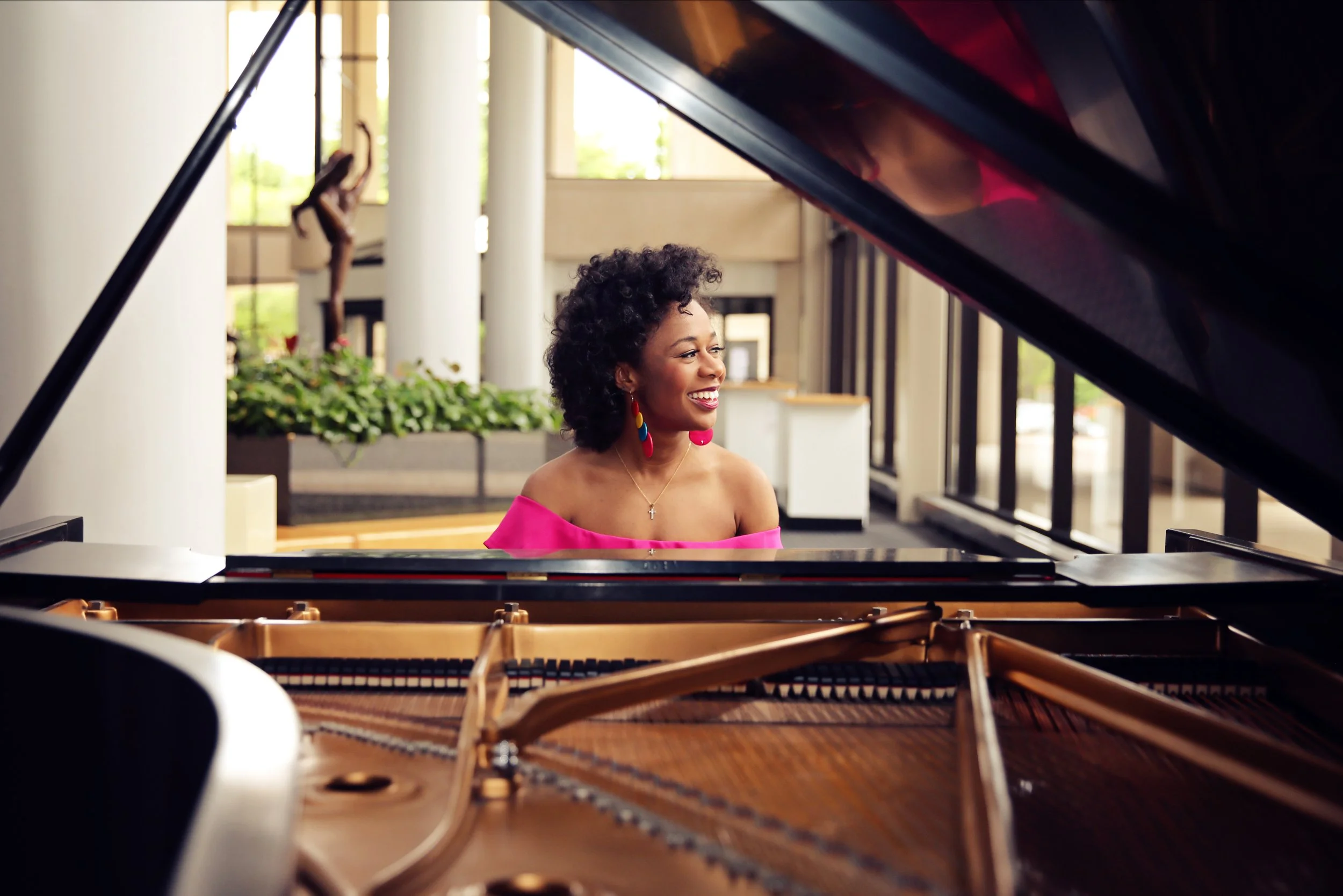 A woman with curly hair smiling while playing a grand piano in a bright indoor space with large windows and decorative plants.