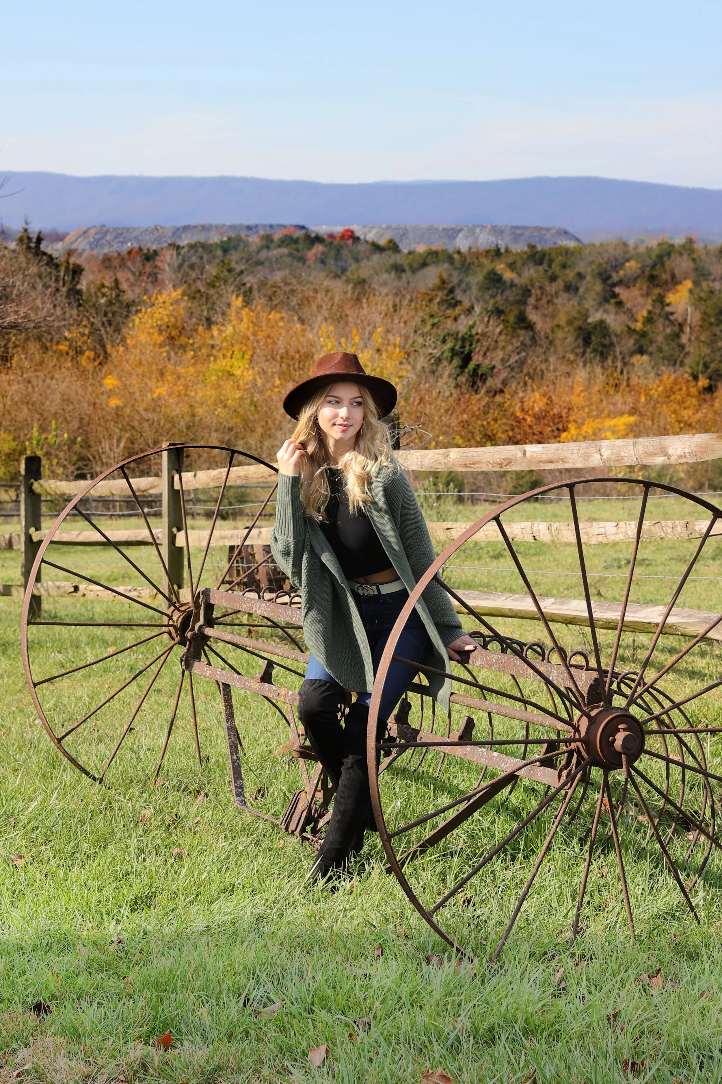 Young woman with blonde hair, wearing a brown hat, green coat, black top, and black boots, sitting on an old rusted horse-drawn plow in a grassy field with trees displaying fall foliage and mountains in the background.