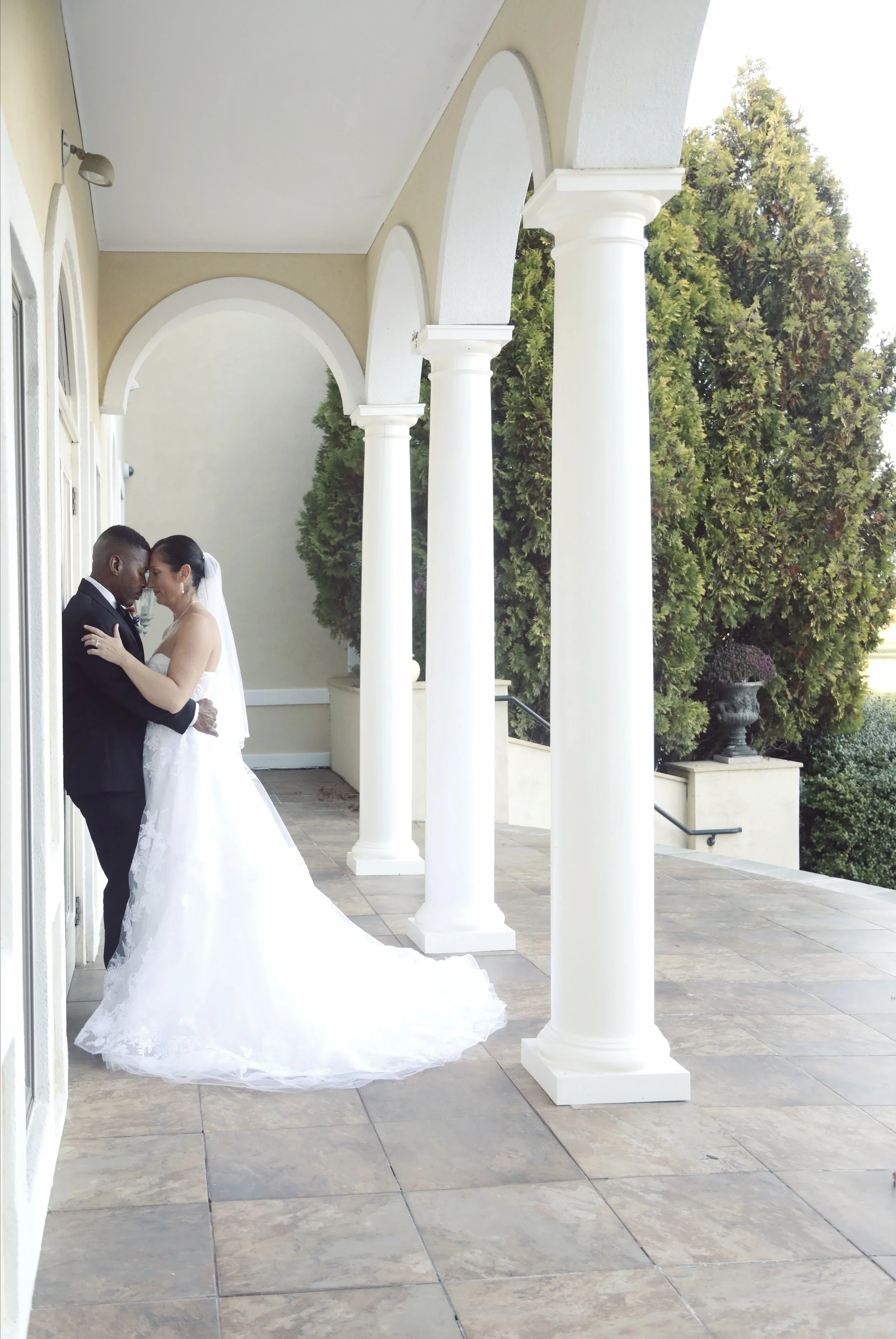 Bride and groom standing closely on a porch with white columns, embracing and touching foreheads, dressed in wedding attire, with greenery and potted plants in the background.