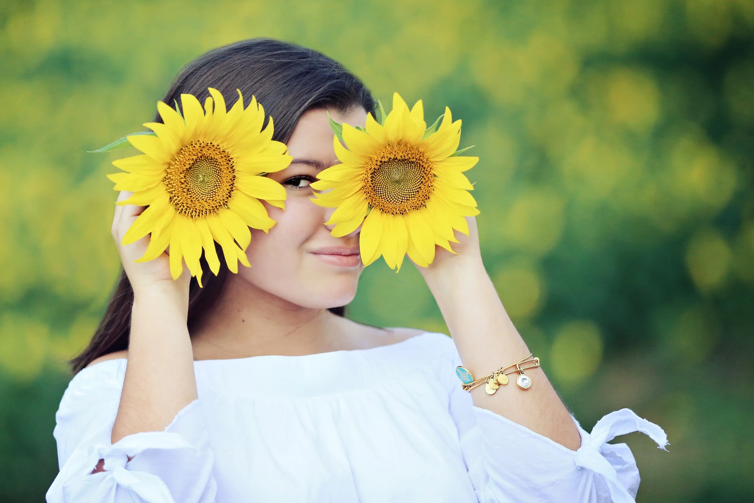 a woman with dark brown hair and light skin holding two large yellow sunflowers in front of her face, partially covering her eyes, outdoors with a green blurred background, wearing a white off-the-shoulder top and a gold bracelet.