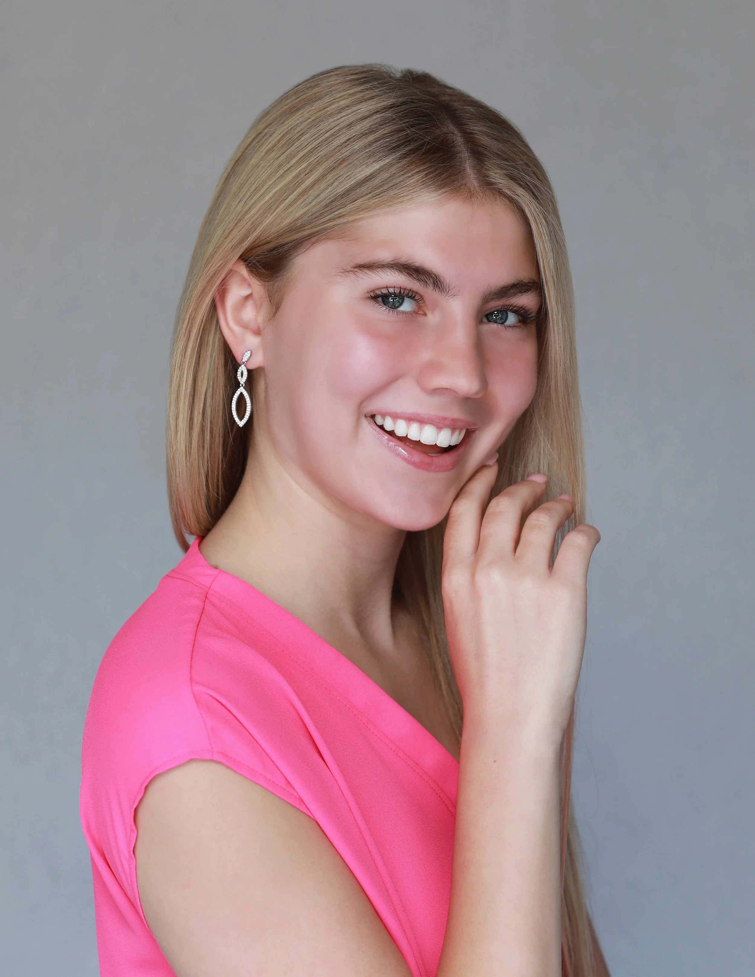 A young woman with long blonde hair, wearing a pink top and silver earrings, smiling and touching her face gently.