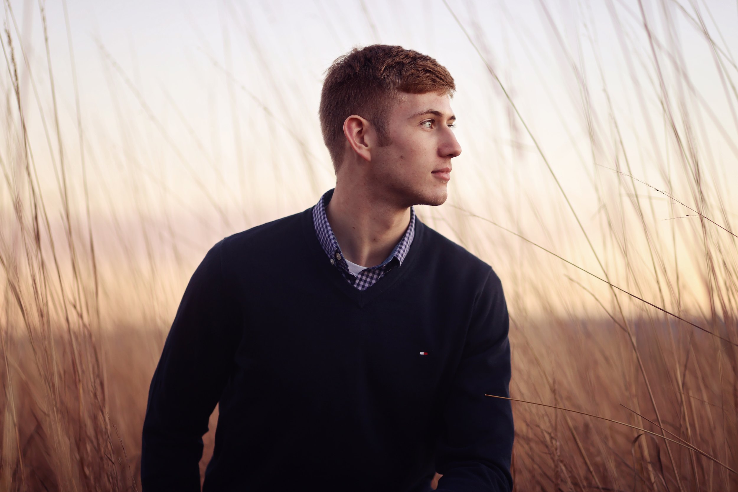 Young man with short brown hair looking to the right, wearing a dark sweater over a checkered shirt, standing outdoors among tall beige grass.