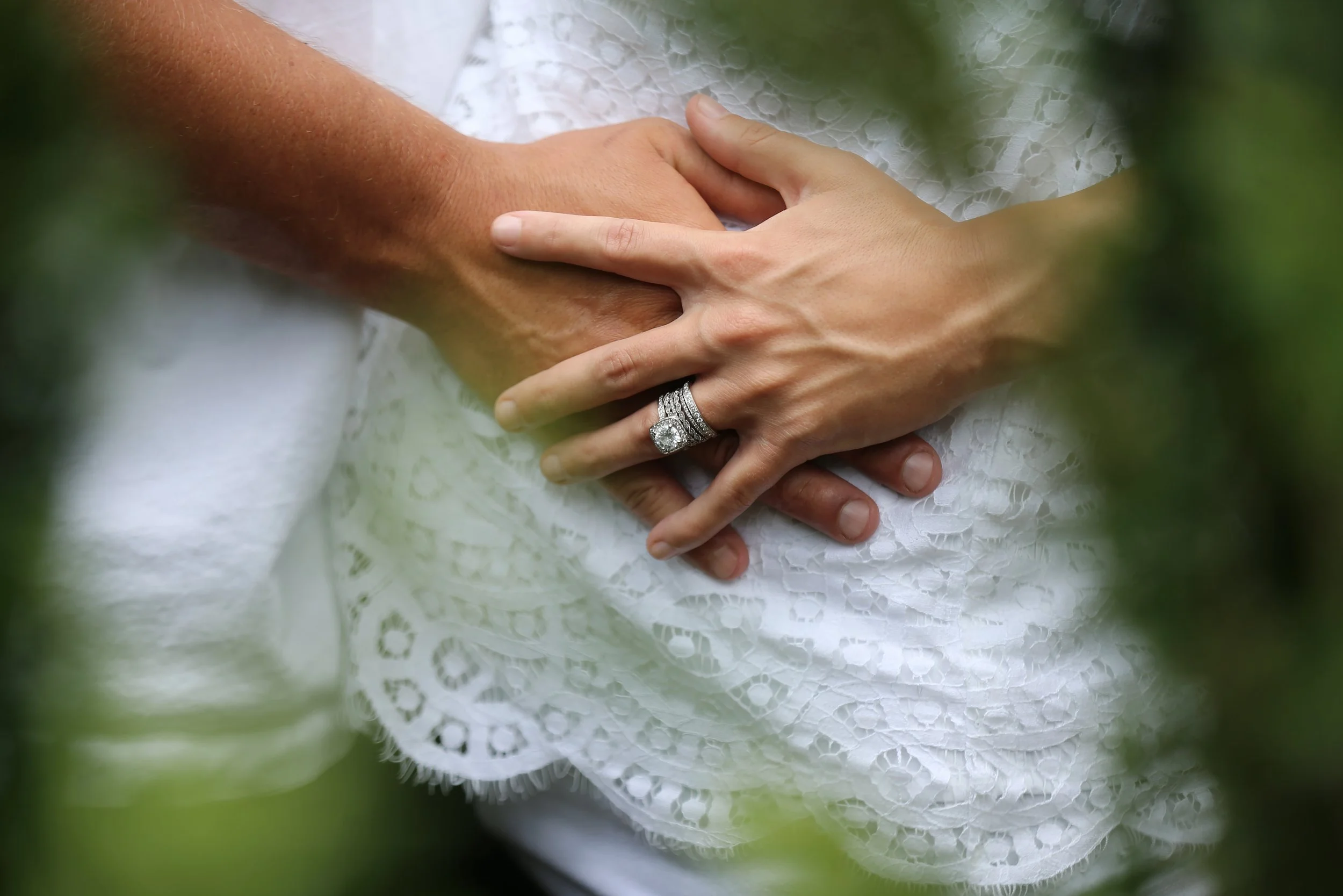 Close-up of a couple holding hands, with the woman's hand resting on top of the man's hand. The woman is wearing an engagement ring and a wedding band with a large central diamond. They are dressed in white lace clothing, possibly wedding attire, and are surrounded by greenery.