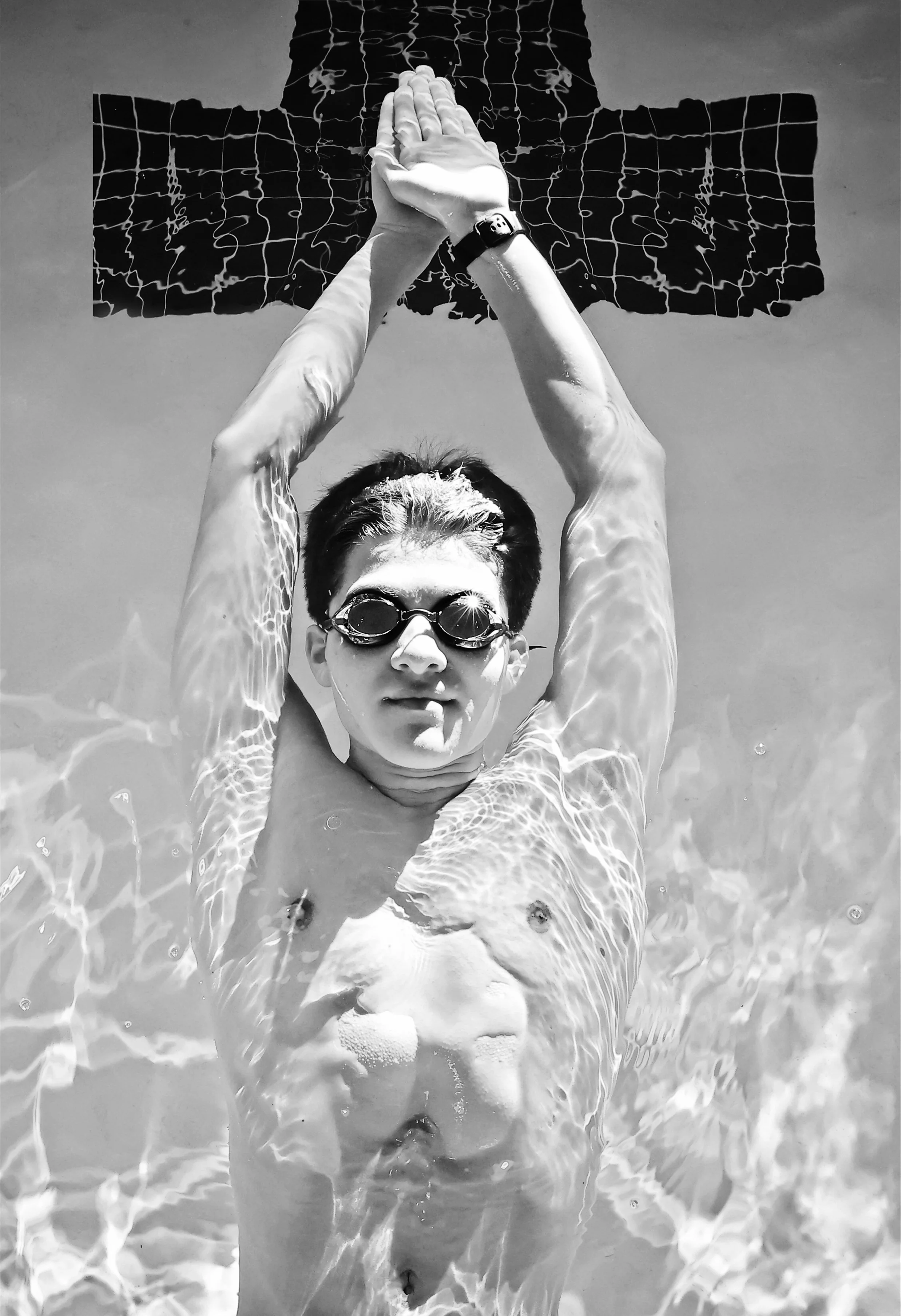 A young man with short dark hair wearing swim goggles and a wristwatch, floating on his back in a swimming pool with his arms stretched above his head. His hands are pressed together and pointed upward, and his face is relaxed. The black-and-white image shows ripples in the water around him and each arm. A large, abstract map or grid design is visible on the ceiling or wall above the pool.