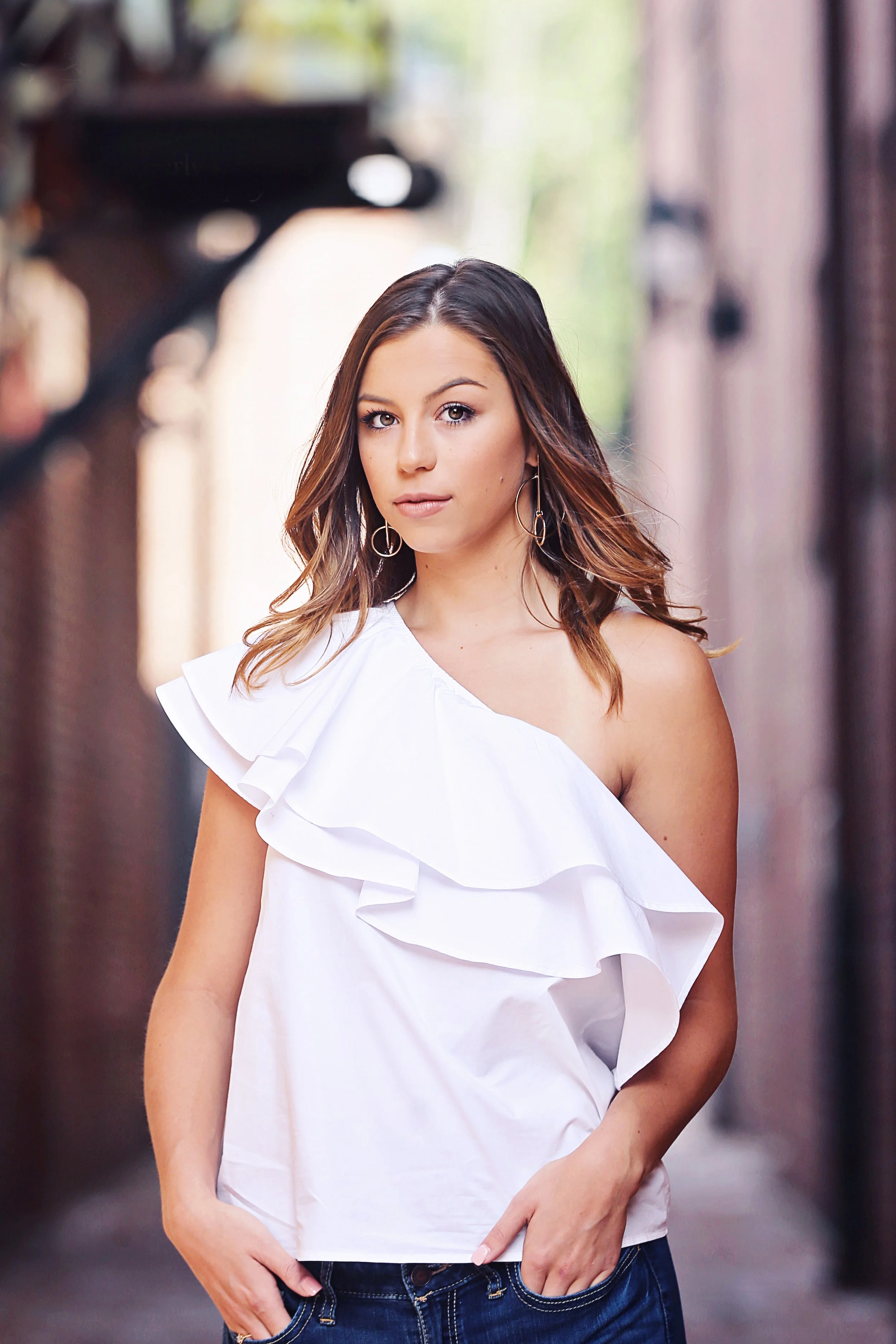 A young woman with shoulder-length brown hair and hoop earrings standing in an alleyway wearing a white one-shoulder ruffled top and jeans.