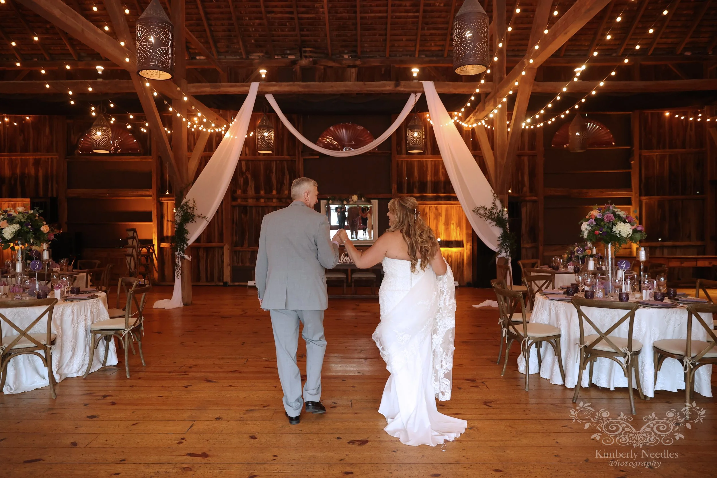 A bride and groom holding hands in a rustic wedding reception venue decorated with string lights, flowers, and fabric drapes.