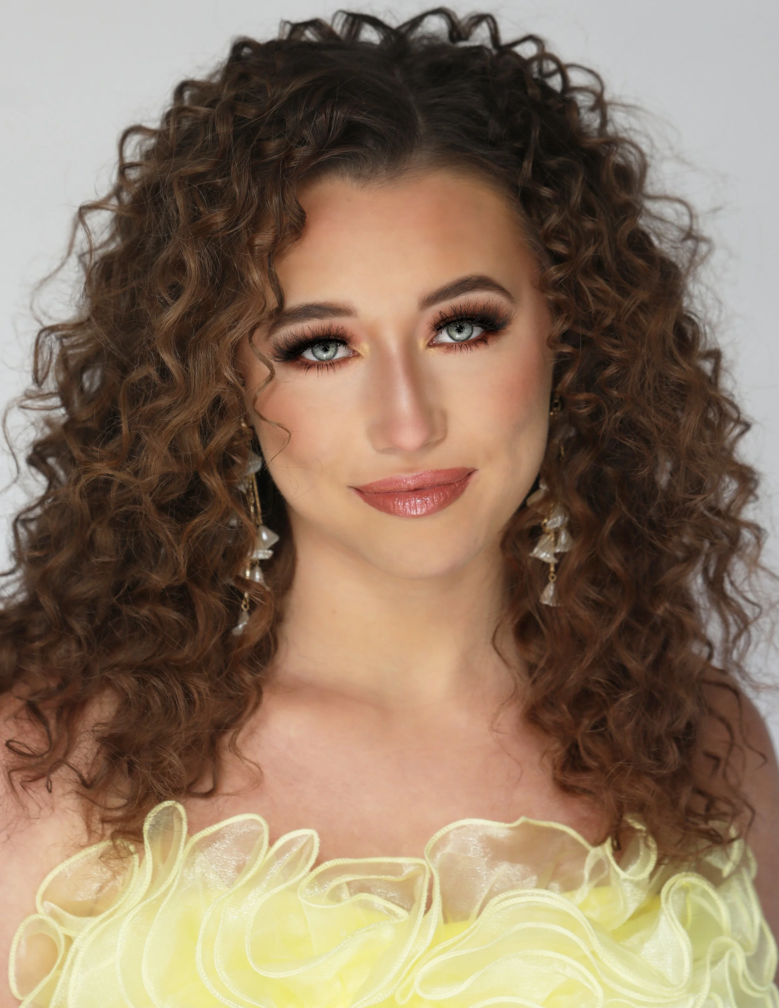 Portrait of a woman with curly brown hair, blue eyes, makeup, and jewelry, wearing a yellow dress with ruffled details.