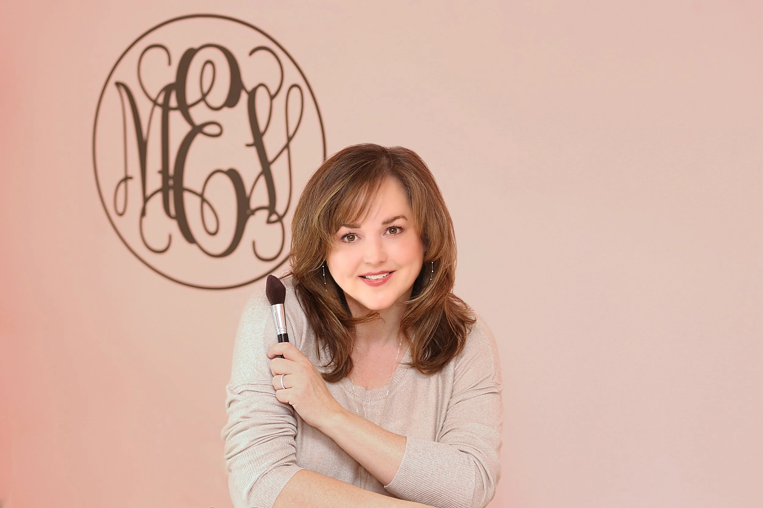 A woman with medium-length brown hair holds a makeup brush and smiles in front of a wall with the name 'Helen Hays' written in script inside a circle.