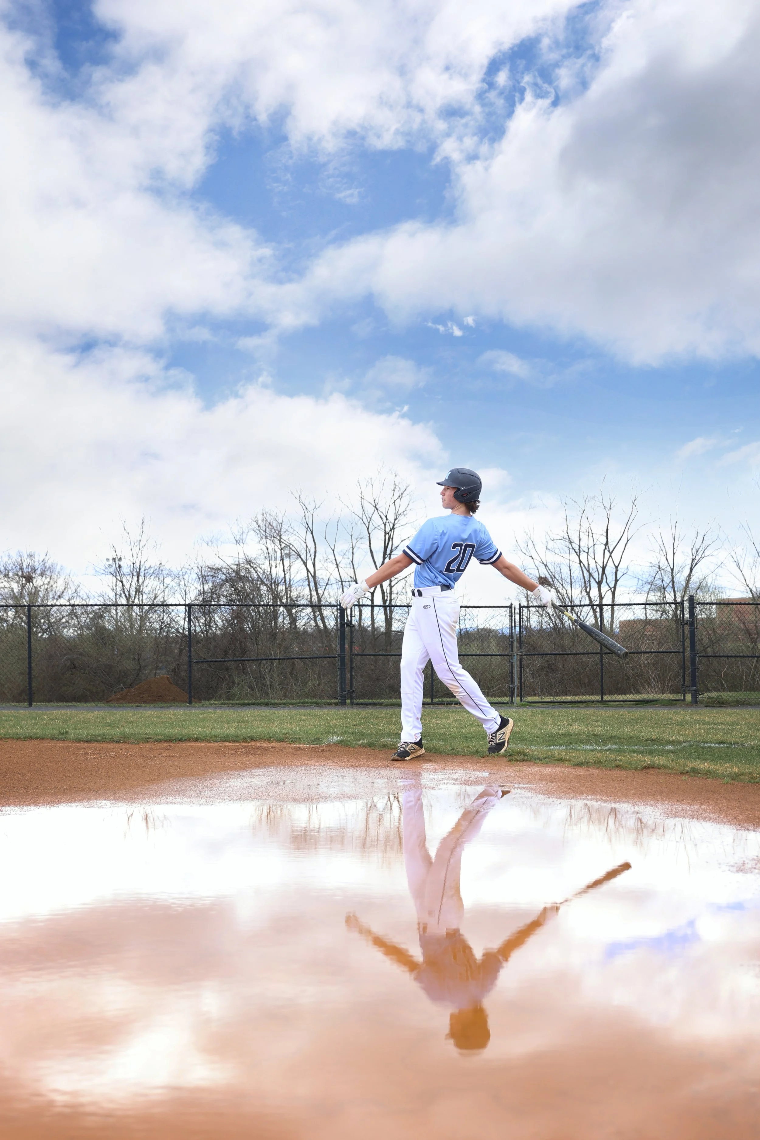 A baseball player in a blue uniform with the number 20 on his back, holding a bat, standing on home plate on a baseball field with a puddle reflecting his image, overcast sky with clouds and bare trees in the background.