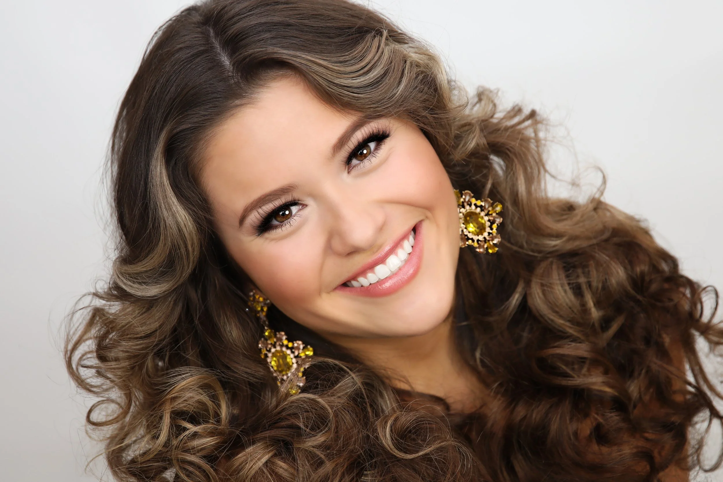 A close-up portrait of a young woman with long, curly brown hair, smiling and showing her teeth, wearing makeup and large gold jewelry with yellow gemstones.