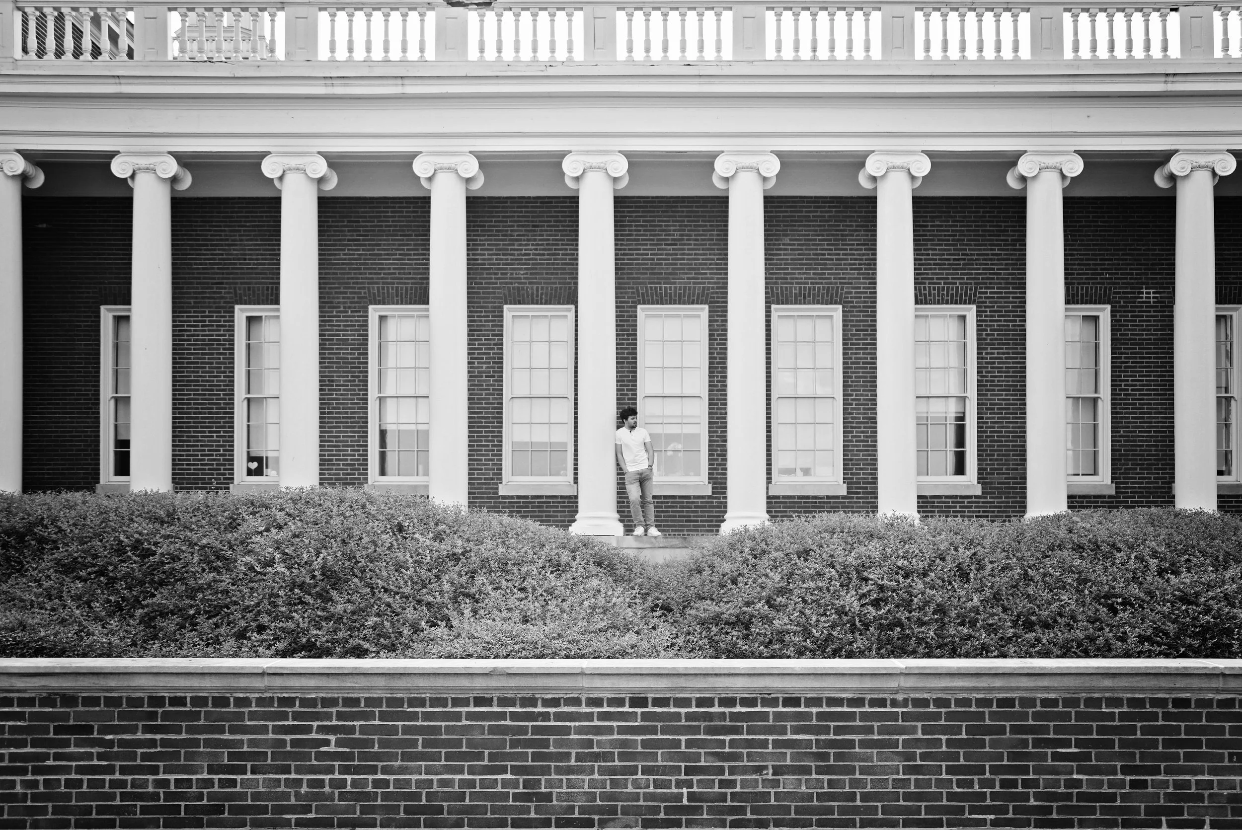 A black and white photo of a person standing on steps in front of a large brick building with tall white columns and multiple windows.