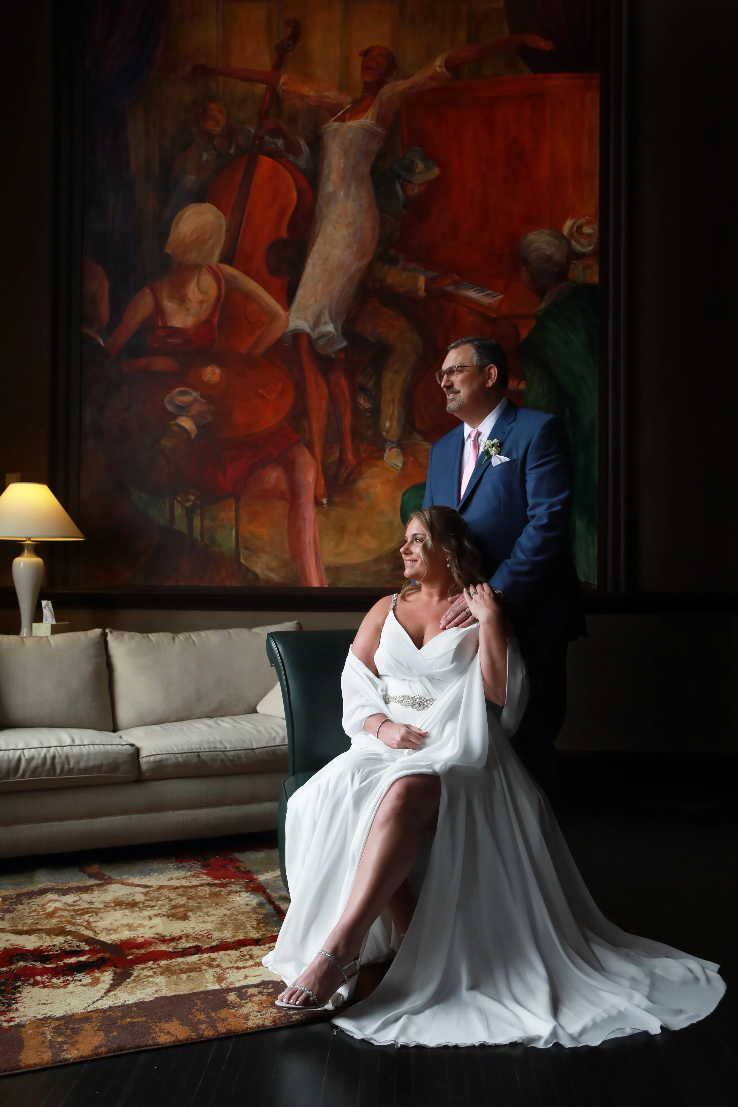 A bride in a white wedding dress sitting on a green chair, with a groom standing behind her, in front of a large painting of musicians and dancers in a dark room.