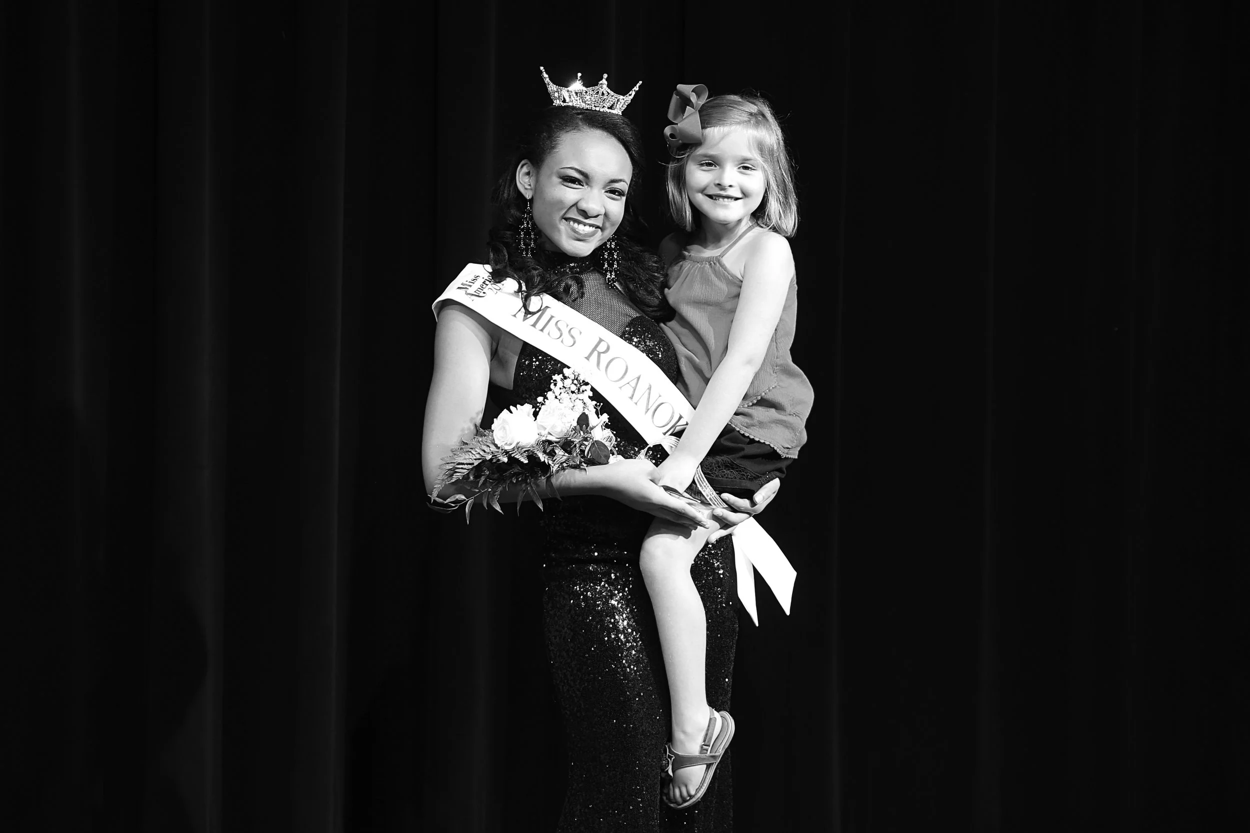 Miss Roanoke wearing a crown and sash, holding a bouquet of flowers, and smiling as she poses with a young girl who is sitting on her arm, both smiling, against a dark background.