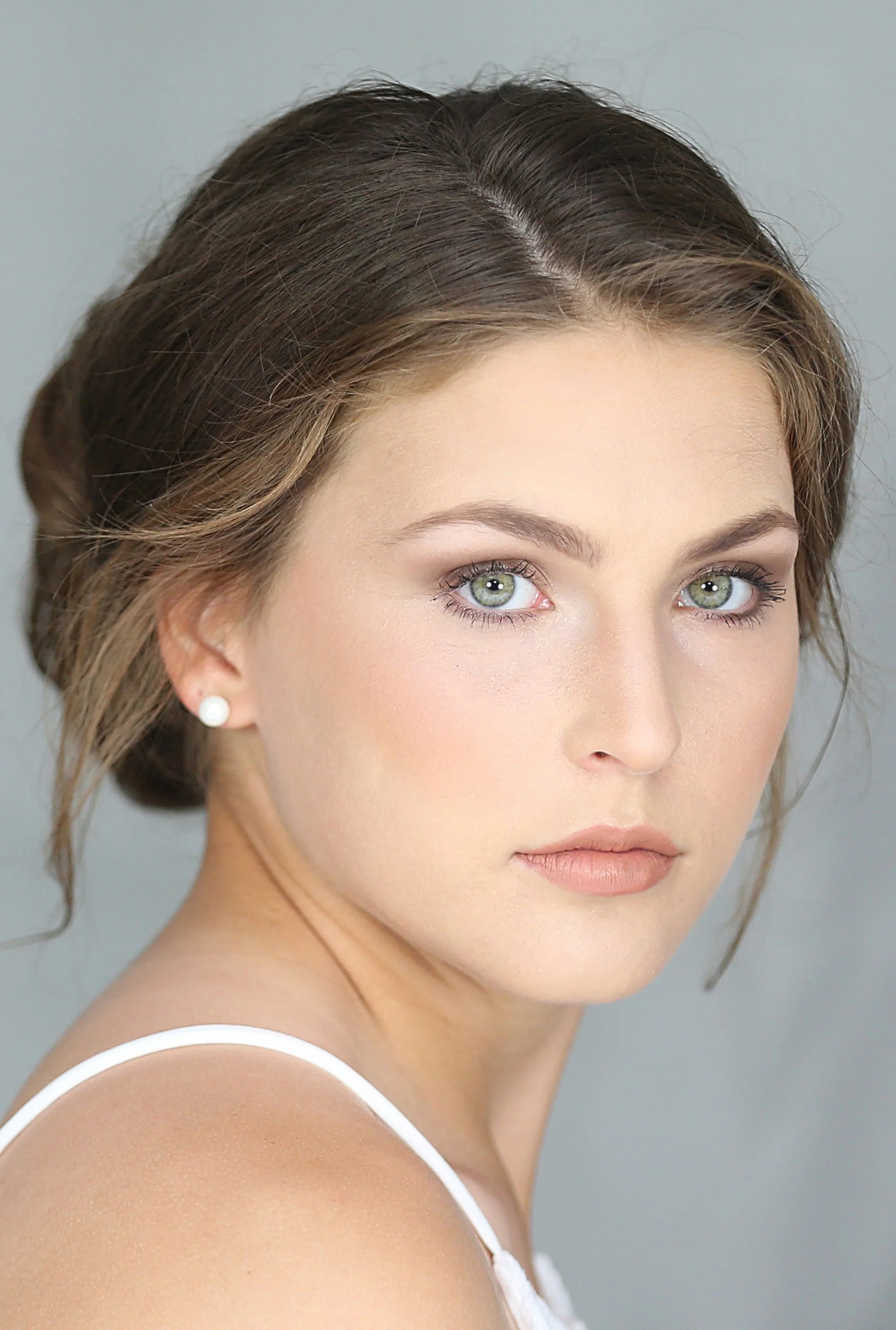 Close-up portrait of a young woman with fair skin, green eyes, light makeup, brown hair styled in an elegant updo, wearing a white top and pearl earrings, against a neutral background.
