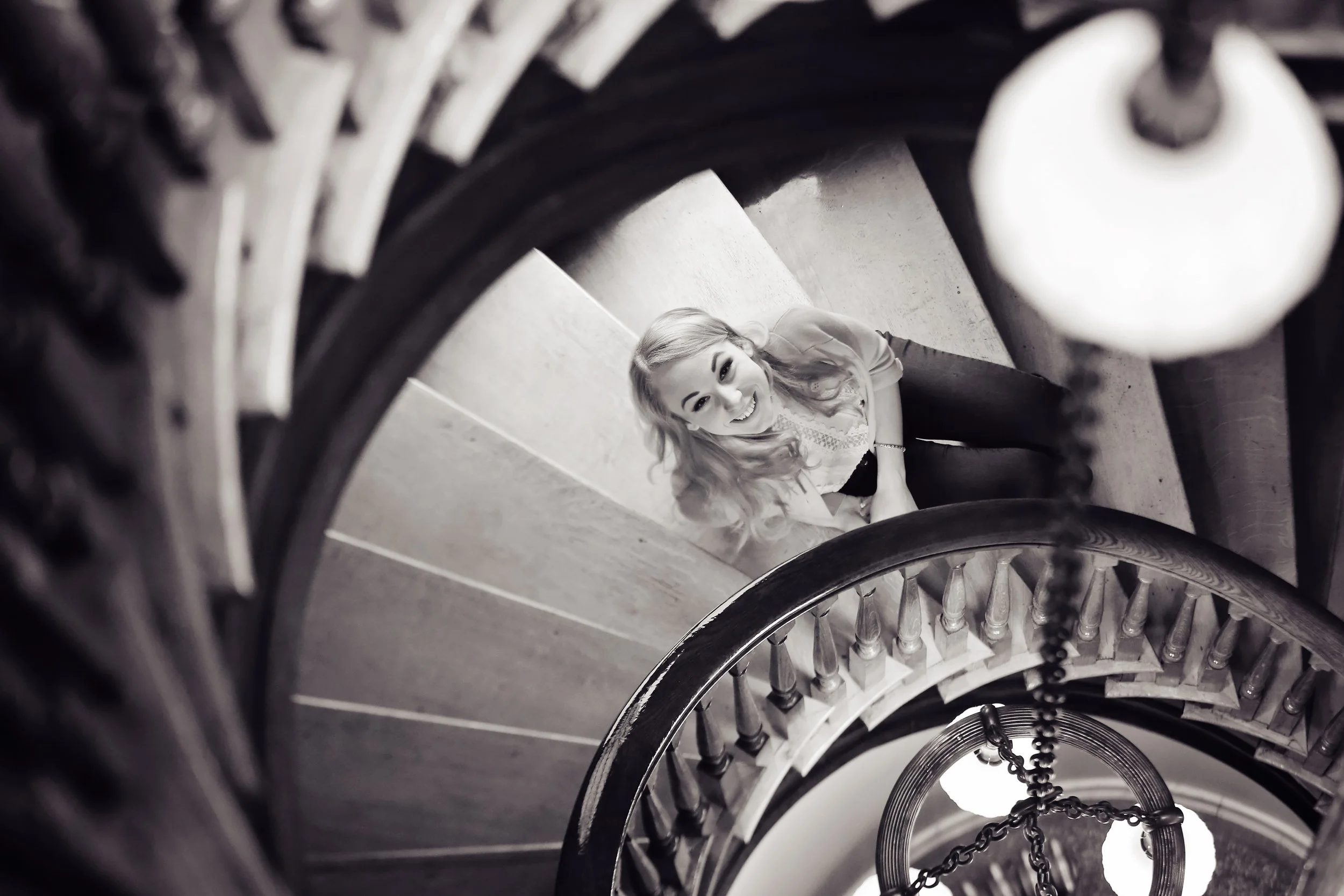 A woman sitting on a staircase at a downward angle, smiling and looking up, photographed from above through the spiral of a staircase railing.