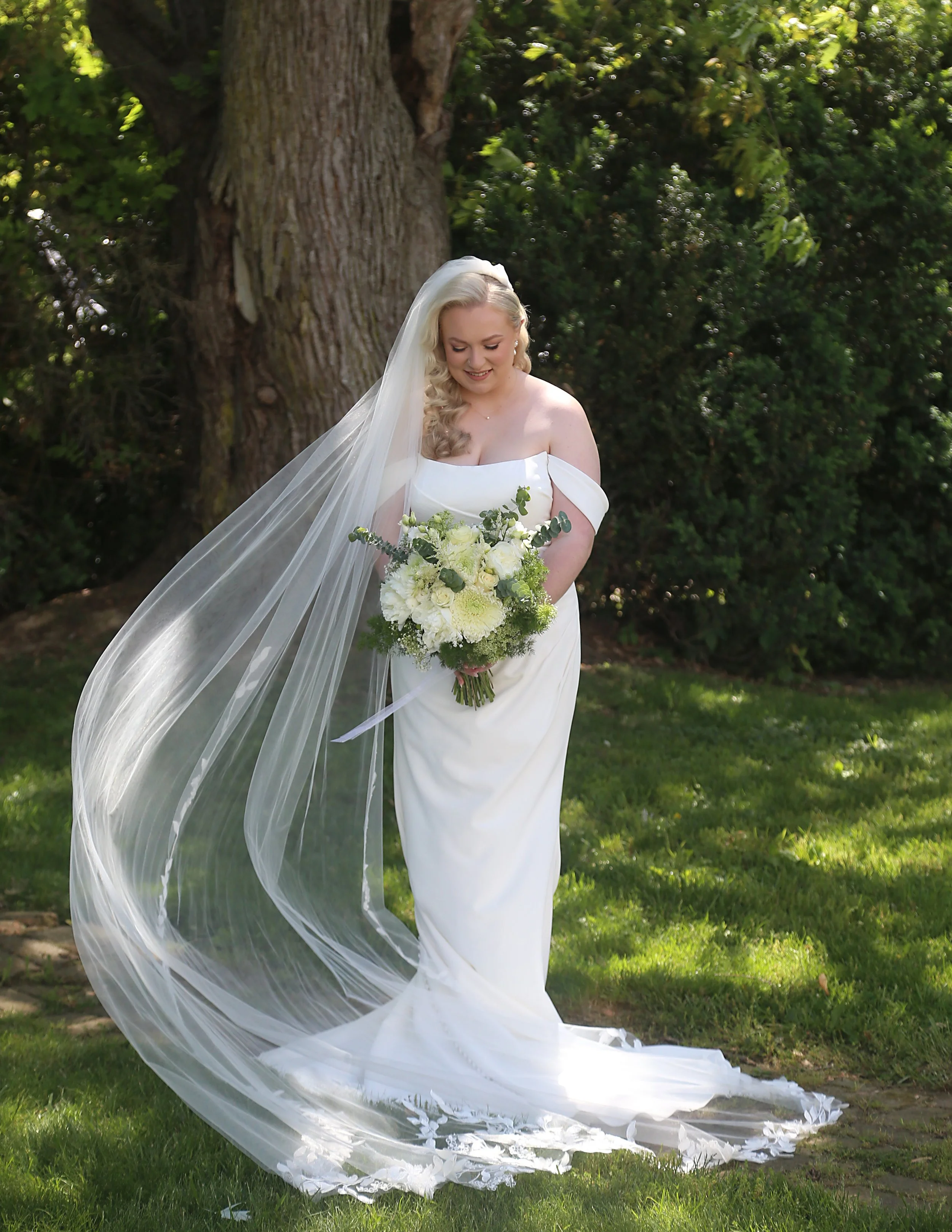 A bride in a white wedding gown standing outdoors near a large tree, holding a bouquet of white and green flowers, with a long veil flowing down her side and smiling.