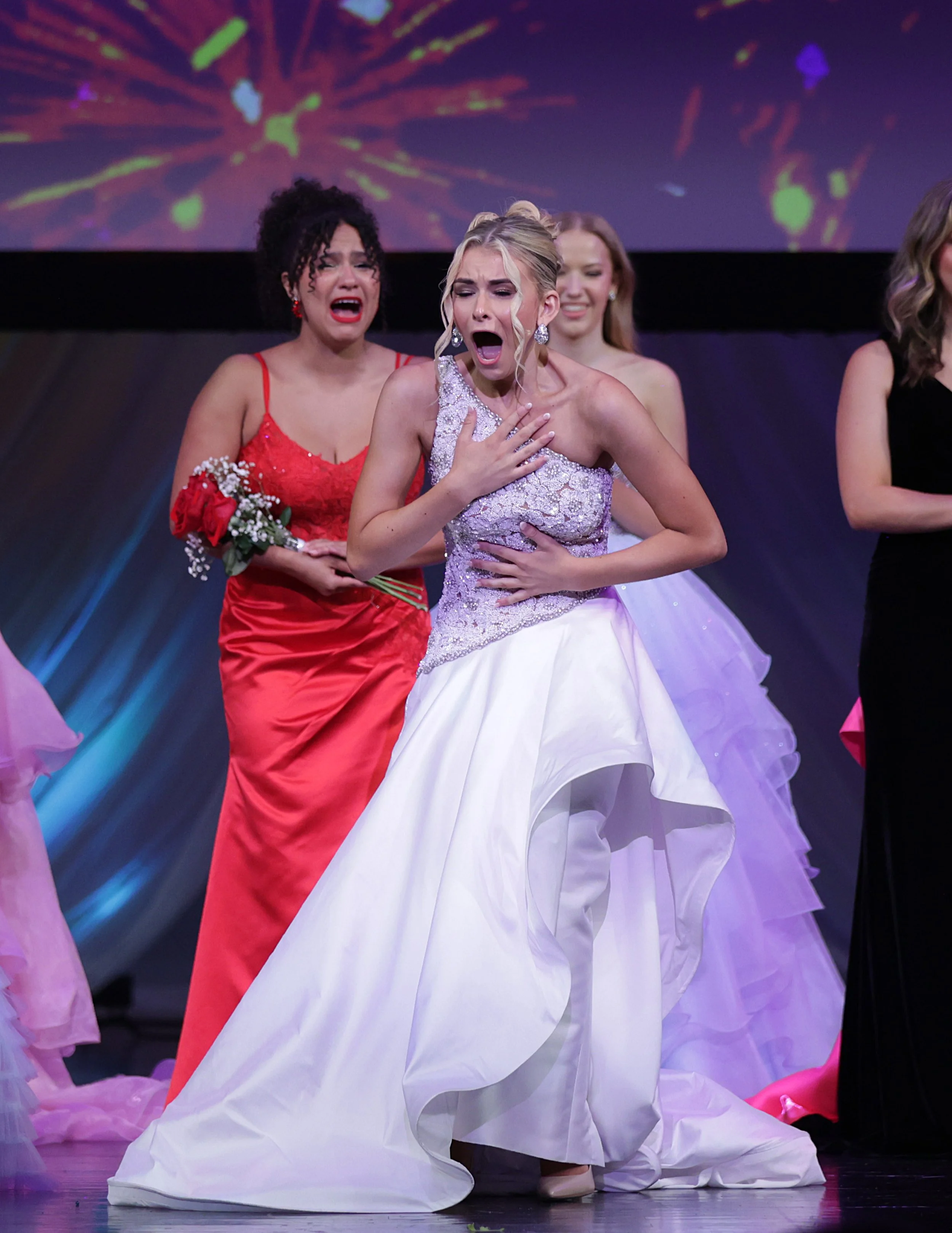 Group of women on stage, one in a white gown, appearing emotional, with others in colorful dresses around her, at a formal event.