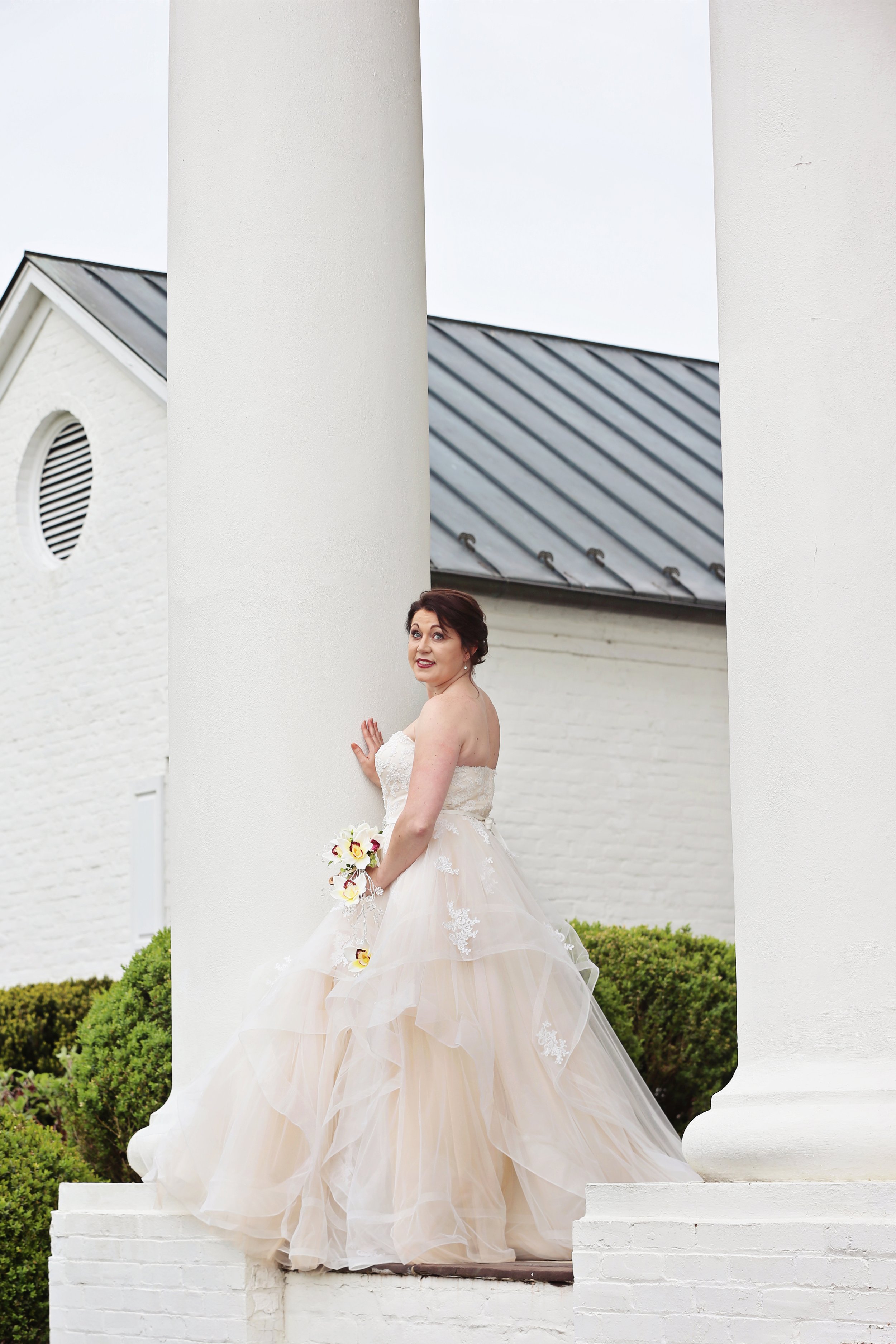 A bride in a strapless wedding dress holding a bouquet of calla lilies stands outdoors near two white columns.