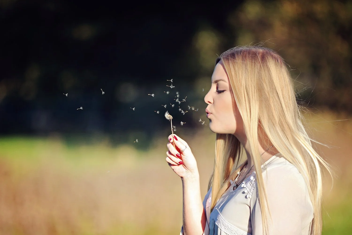 A woman with long blonde hair blowing dandelion seeds outdoors on a sunny day.