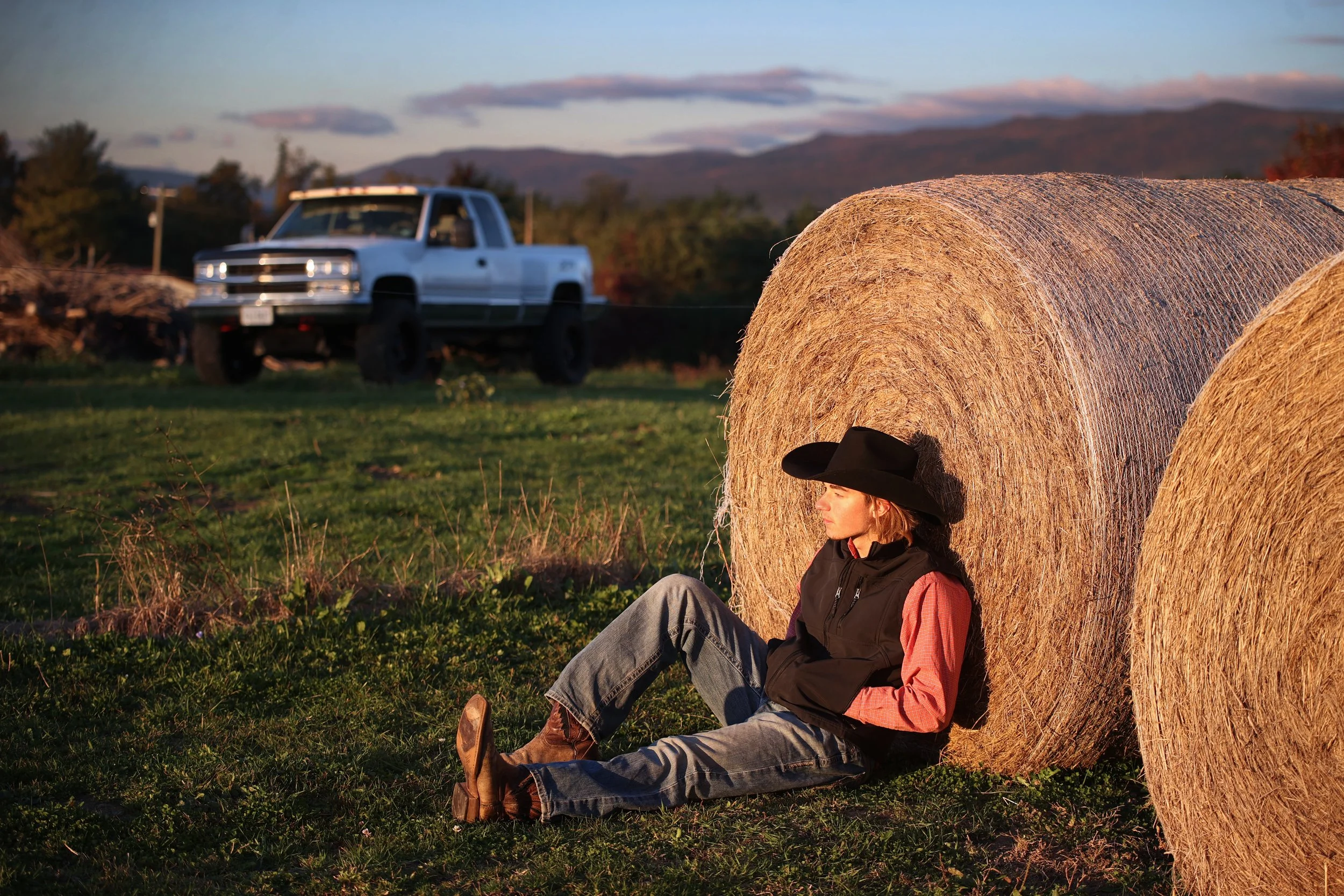 Woman sitting against hay bales in a rural field with a pickup truck and mountains in the background during sunset.