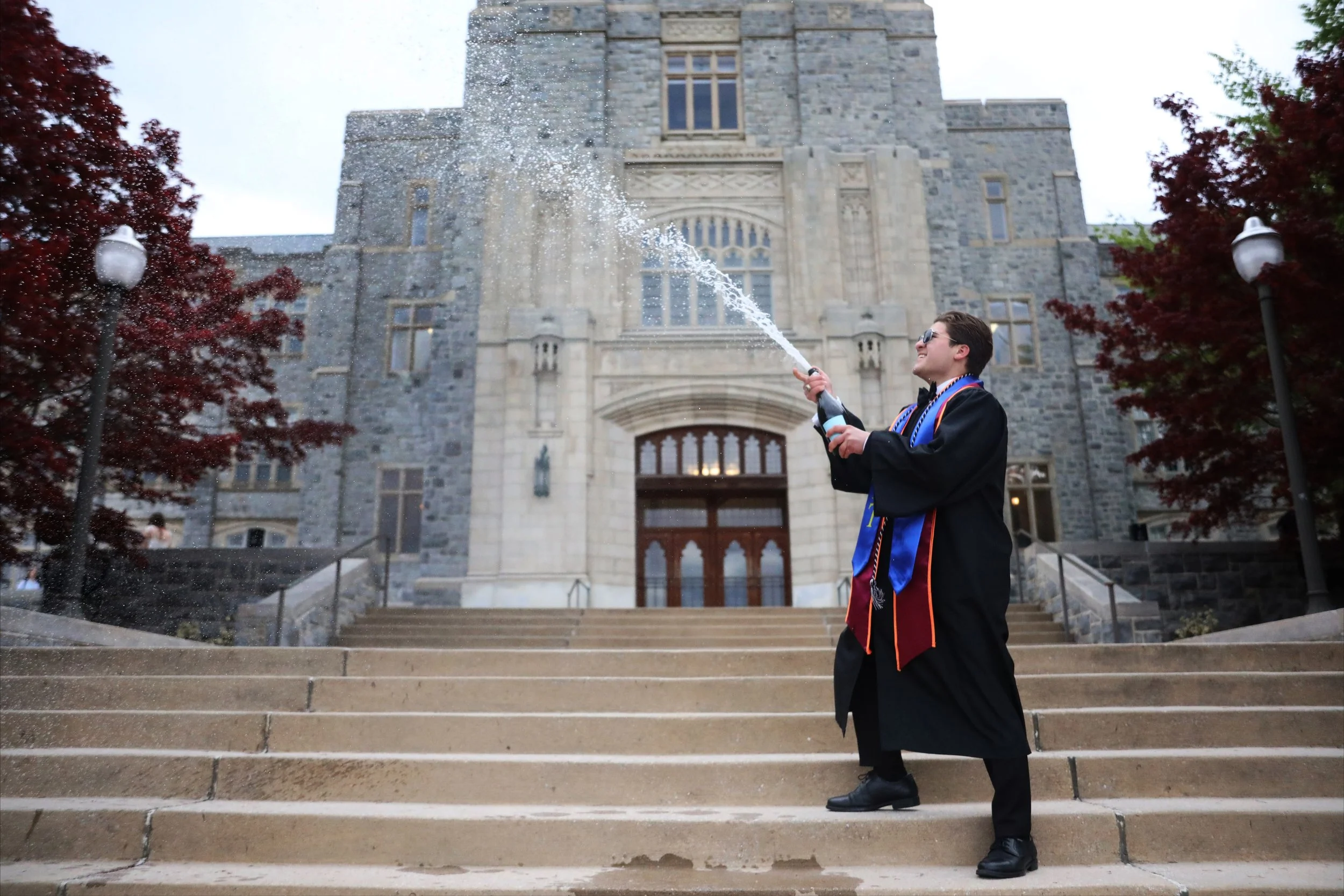 A young man in a graduation gown and cap celebrating on the steps of a historic stone building, spraying champagne.