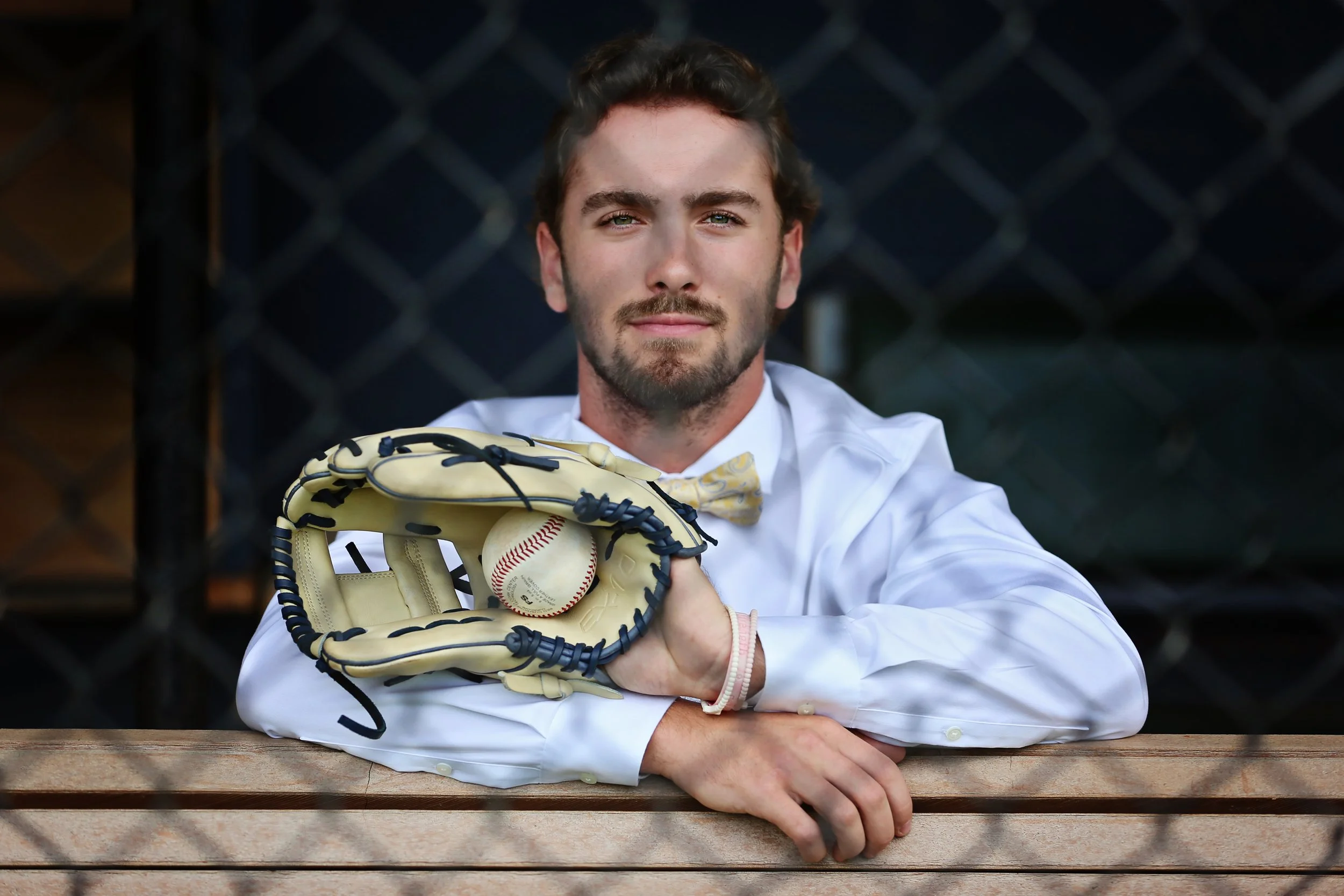 A man in a white dress shirt with a yellow bow tie sitting at a wooden bench, holding a baseball glove with a baseball inside, in front of a chainlink fence.