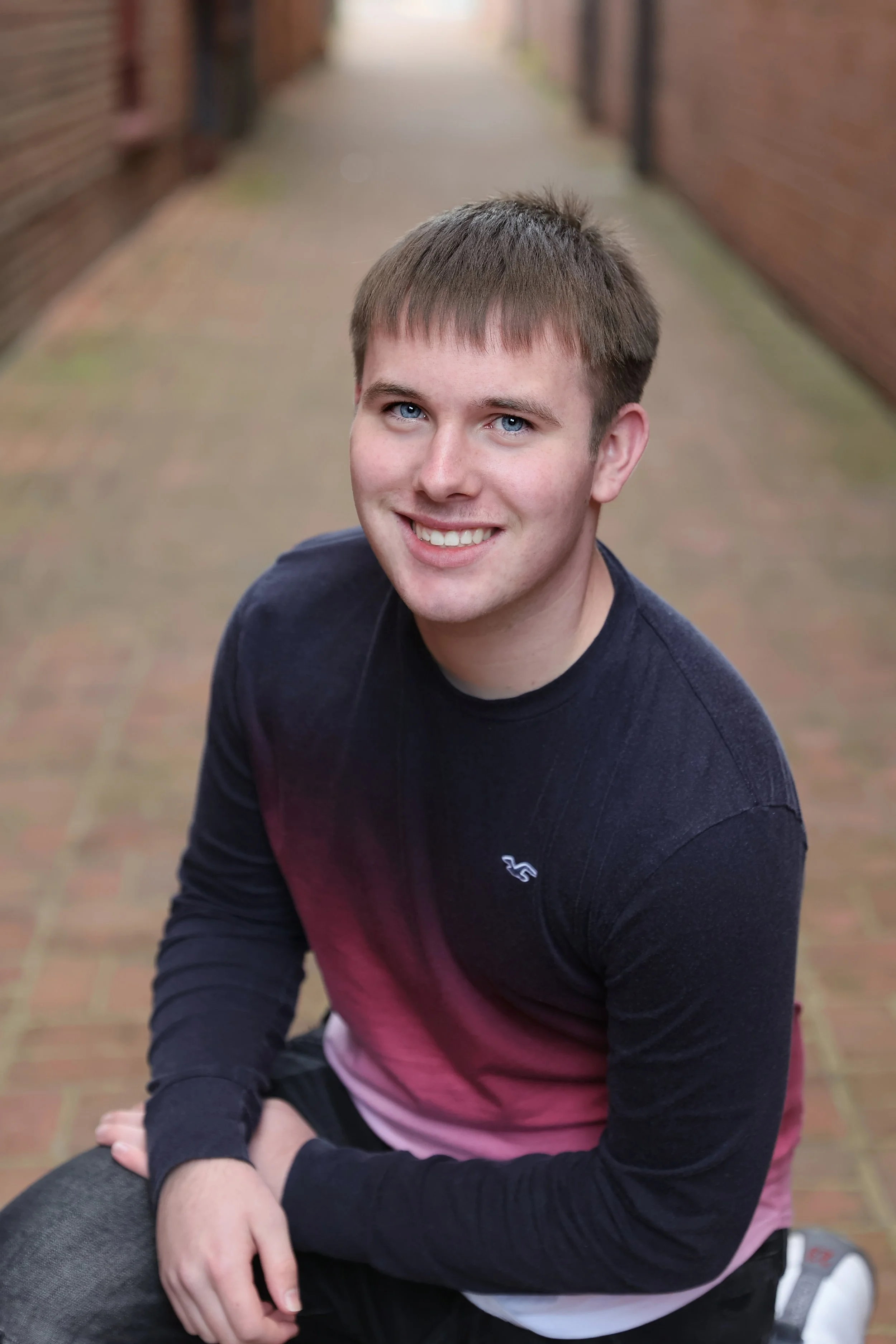 A young man with short light brown hair and blue eyes, smiling, sitting on a brick pathway between brick walls.