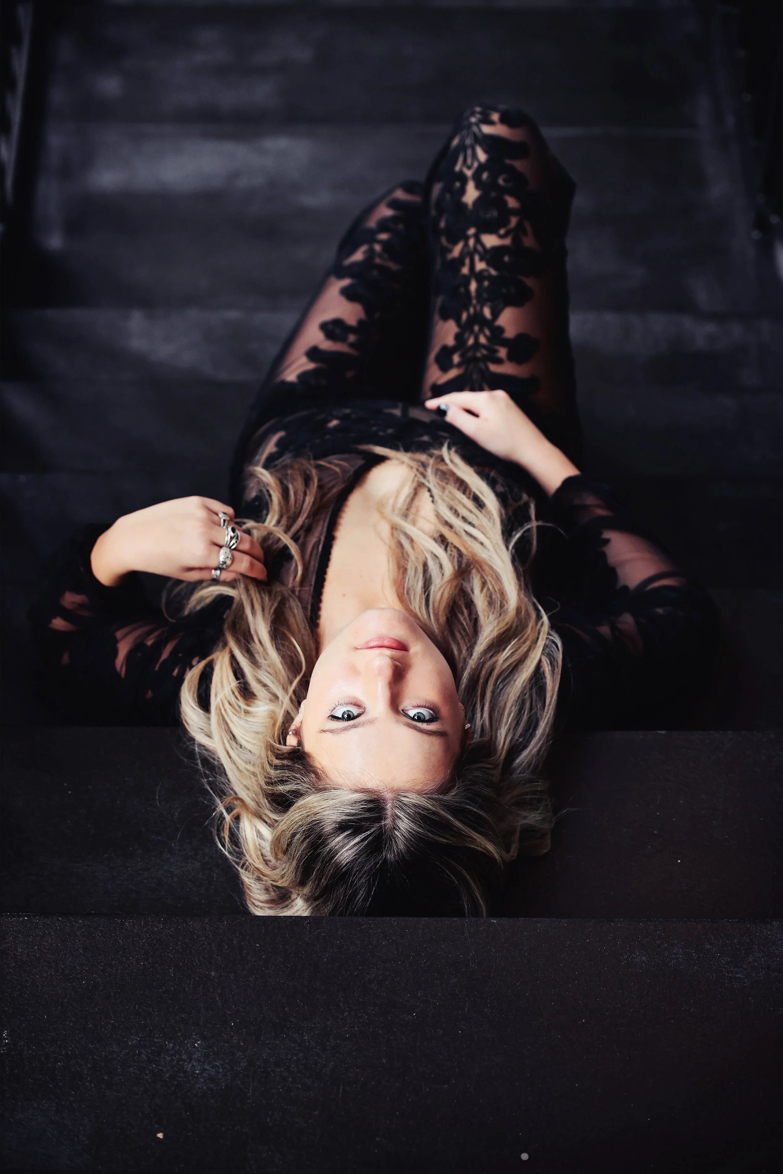 A woman with blonde, wavy hair lying on her back on a black surface, wearing a black lace dress with floral patterns, looking up at the camera.