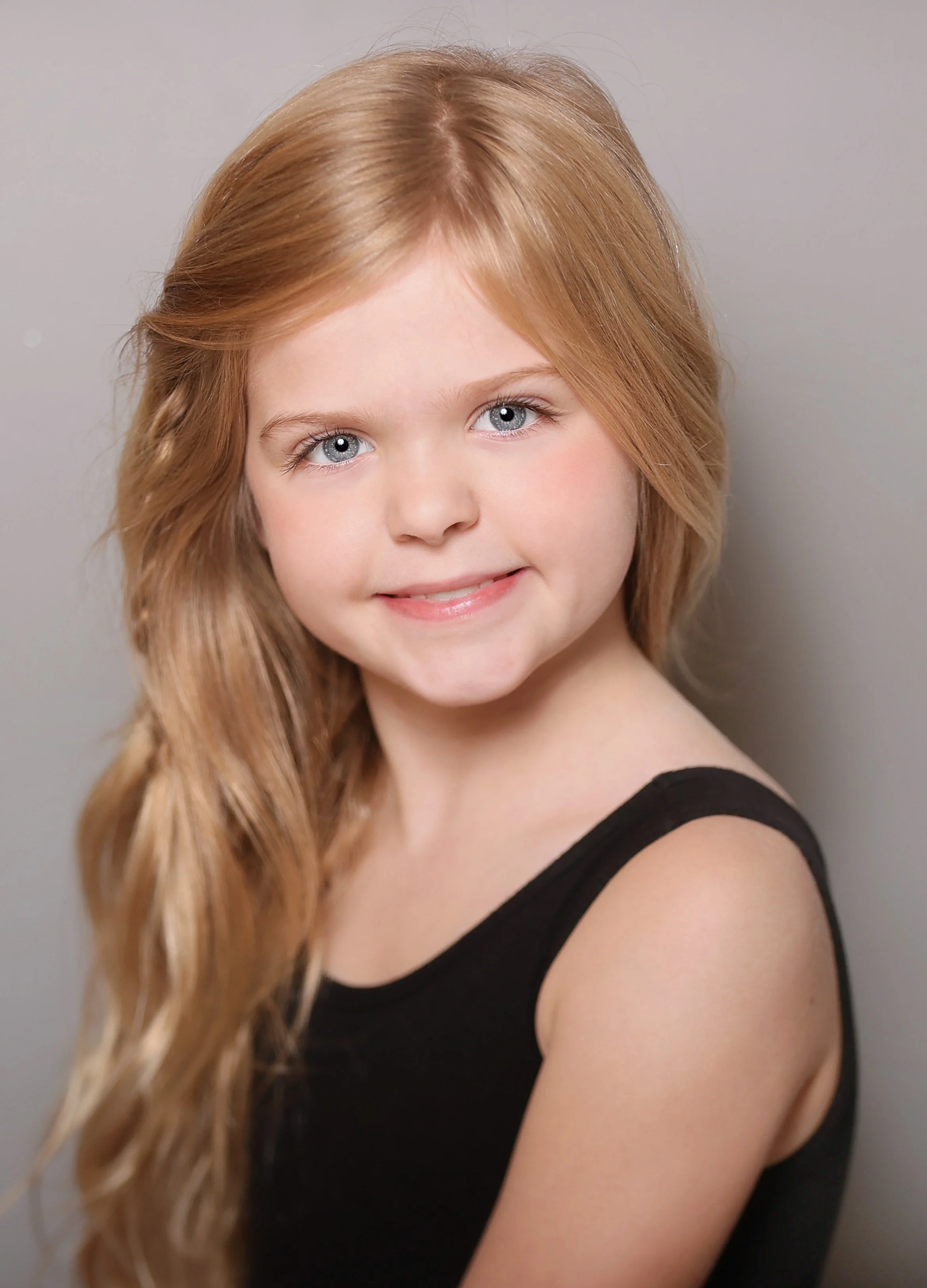 Close-up of a young girl with long, wavy red hair and blue eyes, smiling and wearing a black sleeveless top, against a neutral background.