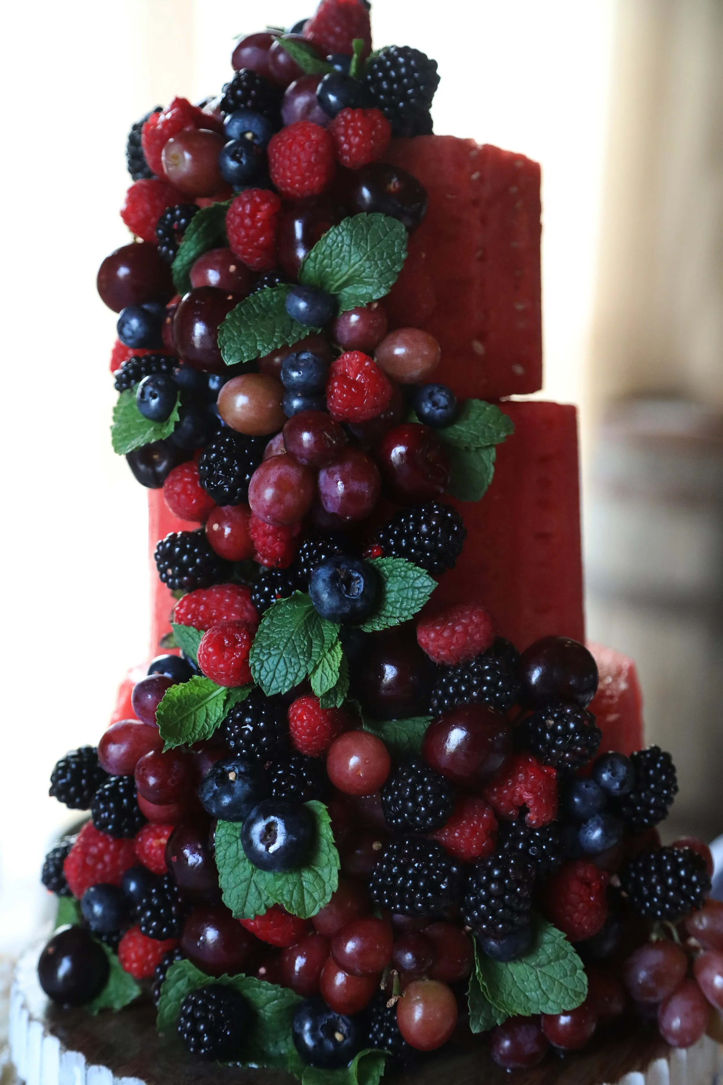 A tall cake decorated with an assortment of mixed berries, including blueberries, raspberries, blackberries, and red grapes, topped with fresh green mint leaves.