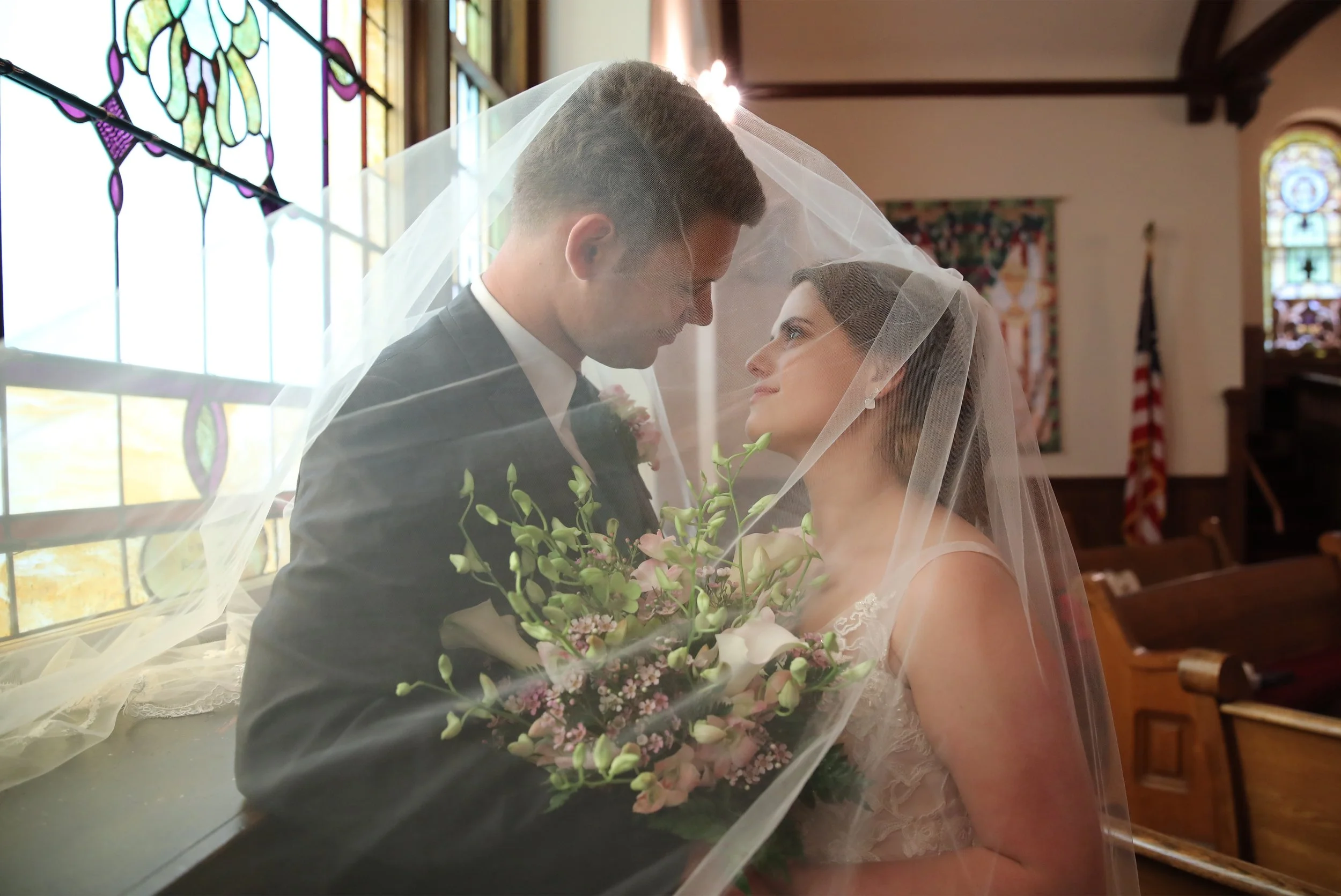 A bride and groom standing close together under a veil inside a church, holding a bouquet of pink and white flowers, with stained glass windows and an American flag in the background.