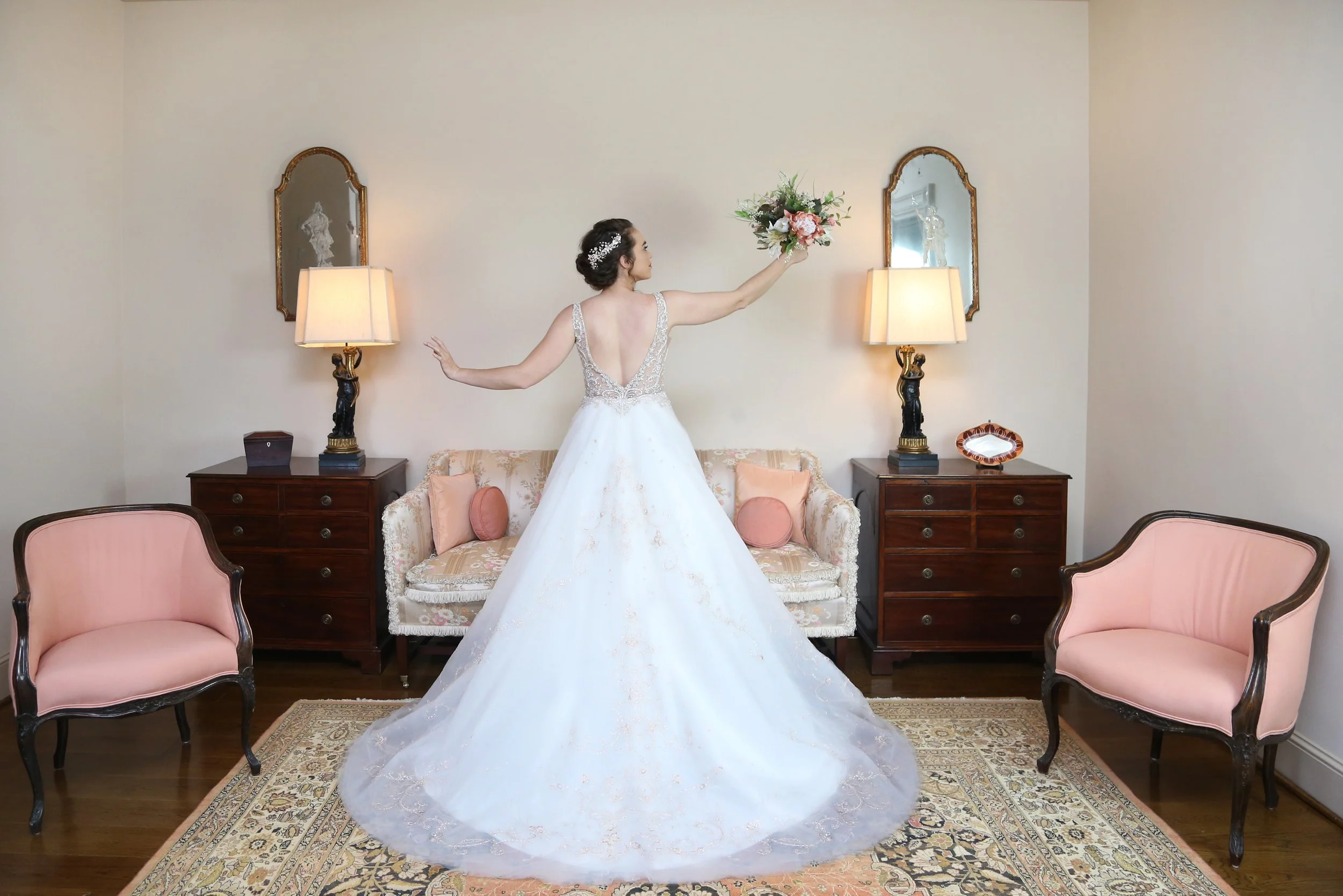 A bride in a wedding dress standing in a vintage-style living room, holding a bouquet of flowers, with two pink armchairs, a floral couch, and wooden side tables with lamps and mirrors on the wall.