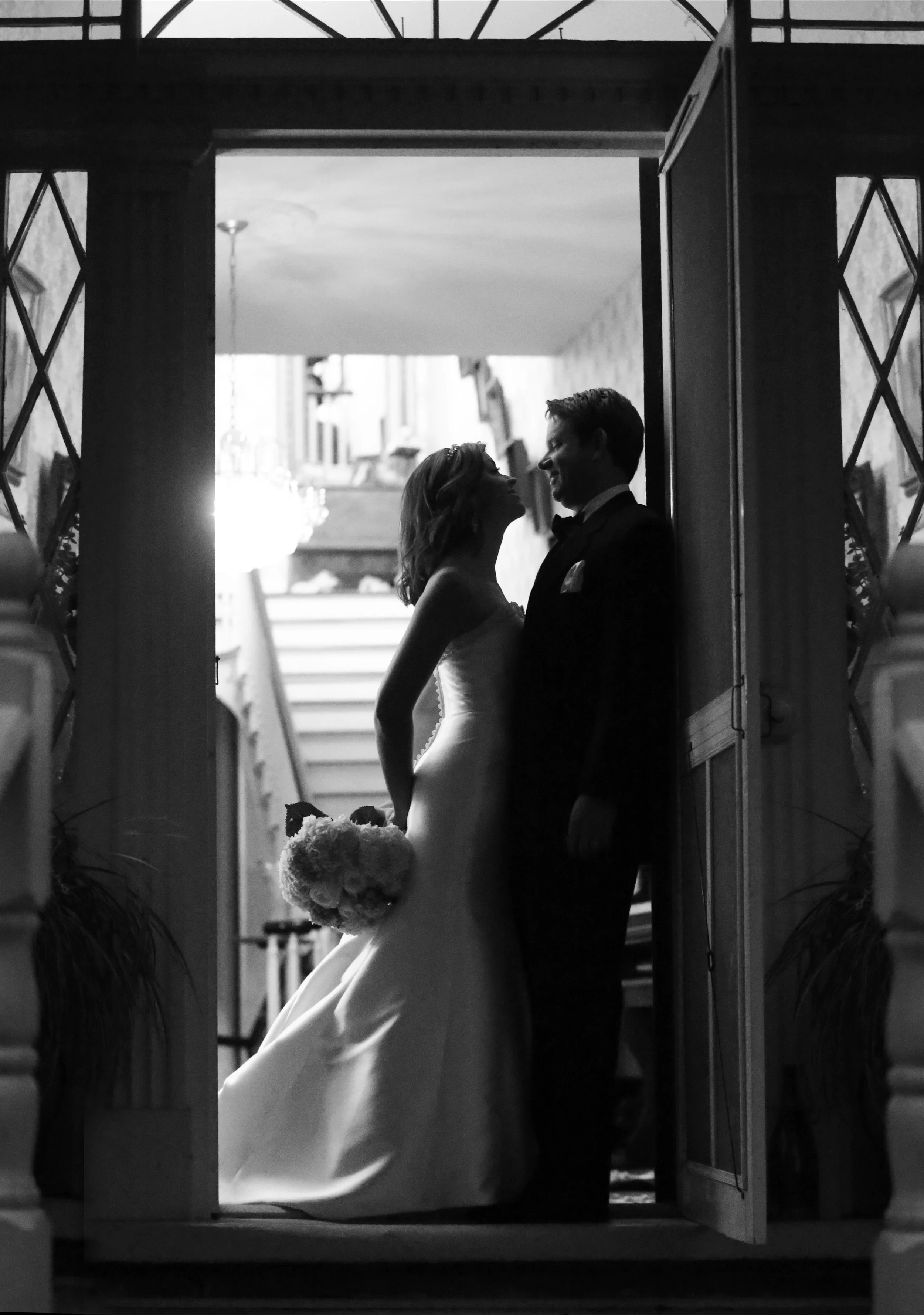 Black and white photo of a bride and groom standing close together at their wedding, inside a doorway, with the bride holding a bouquet of flowers.