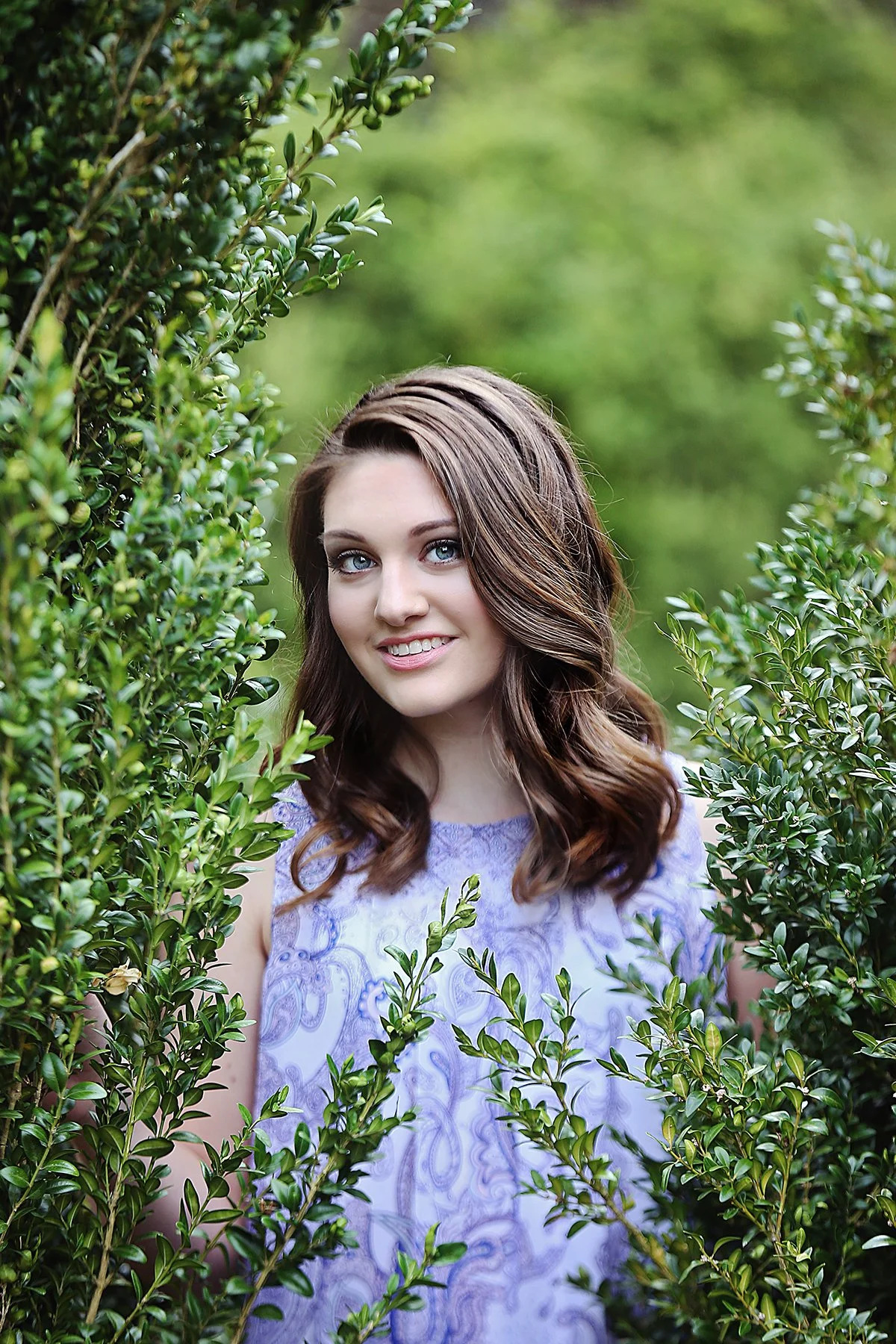 A young woman with long, wavy brown hair and blue eyes smiling while standing in a lush green garden surrounded by dense bushes.