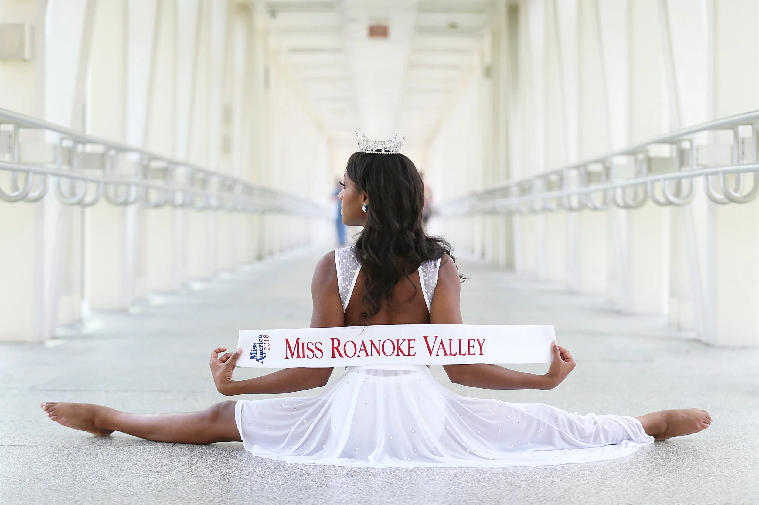 A woman in a white dress and crown sitting on the floor in a hallway performing a front split, holding a sash that reads 'Miss Roanoke Valley,' with a view of the corridor extending into the distance.